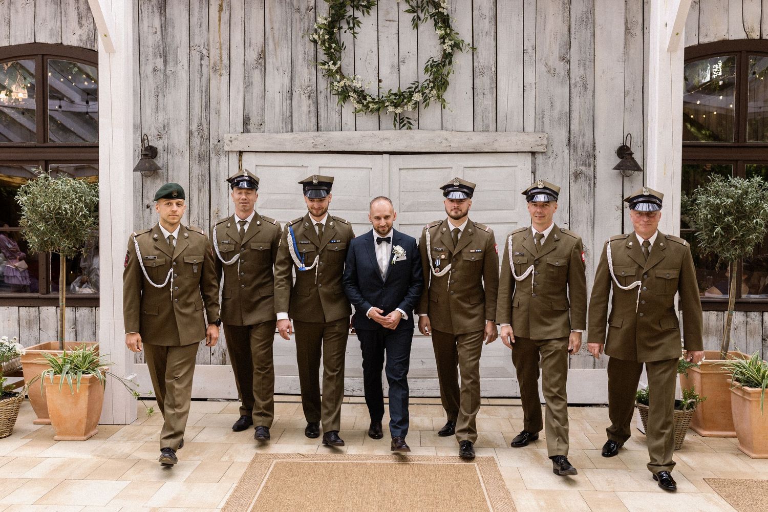 Military personnel in dress uniforms stand together outside a rustic barn entrance with a decorative wreath above.