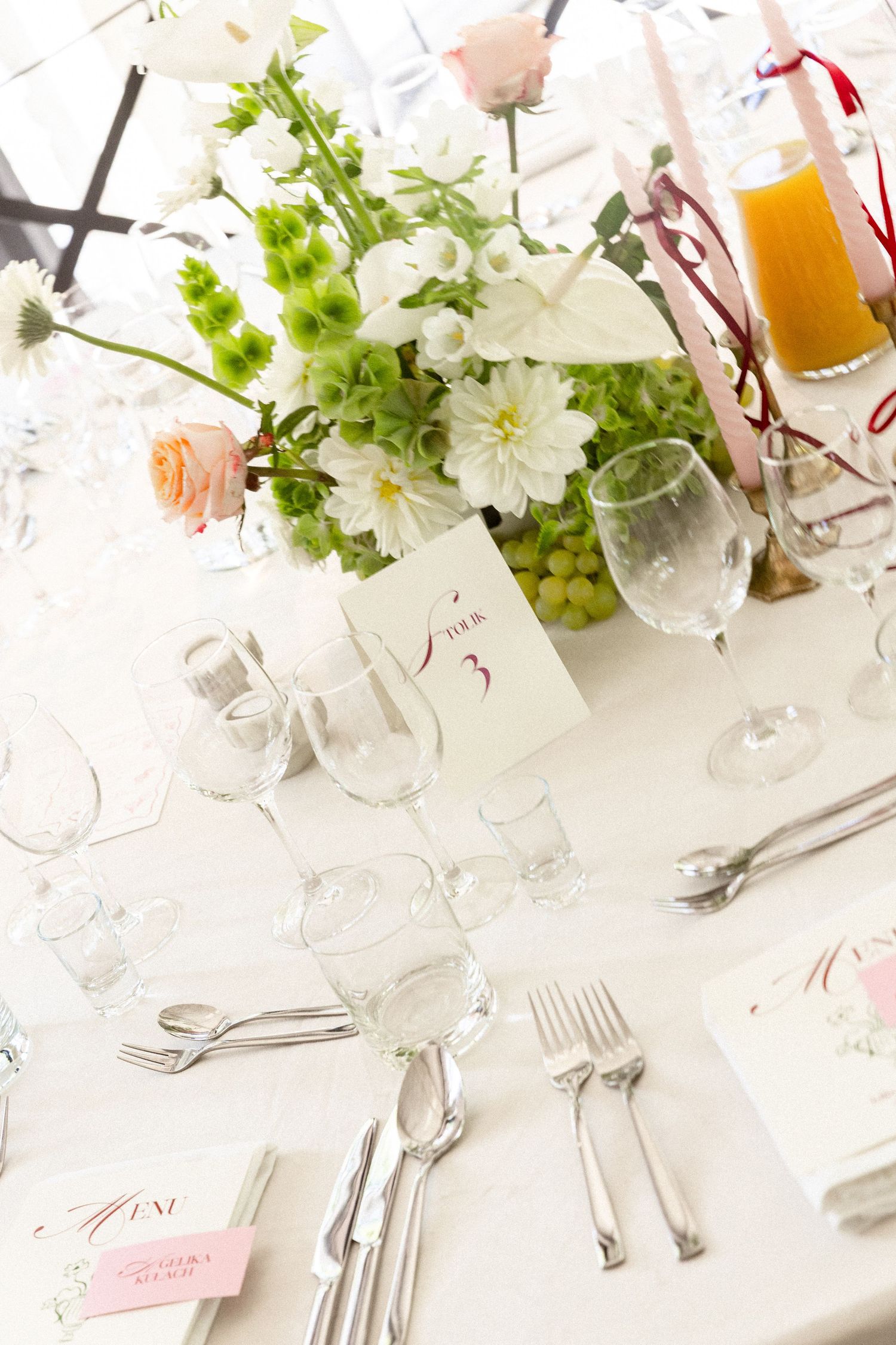 An elegant wedding table setting with white flowers, pink roses, cutlery, wine glasses and orange juice in glass bottles.