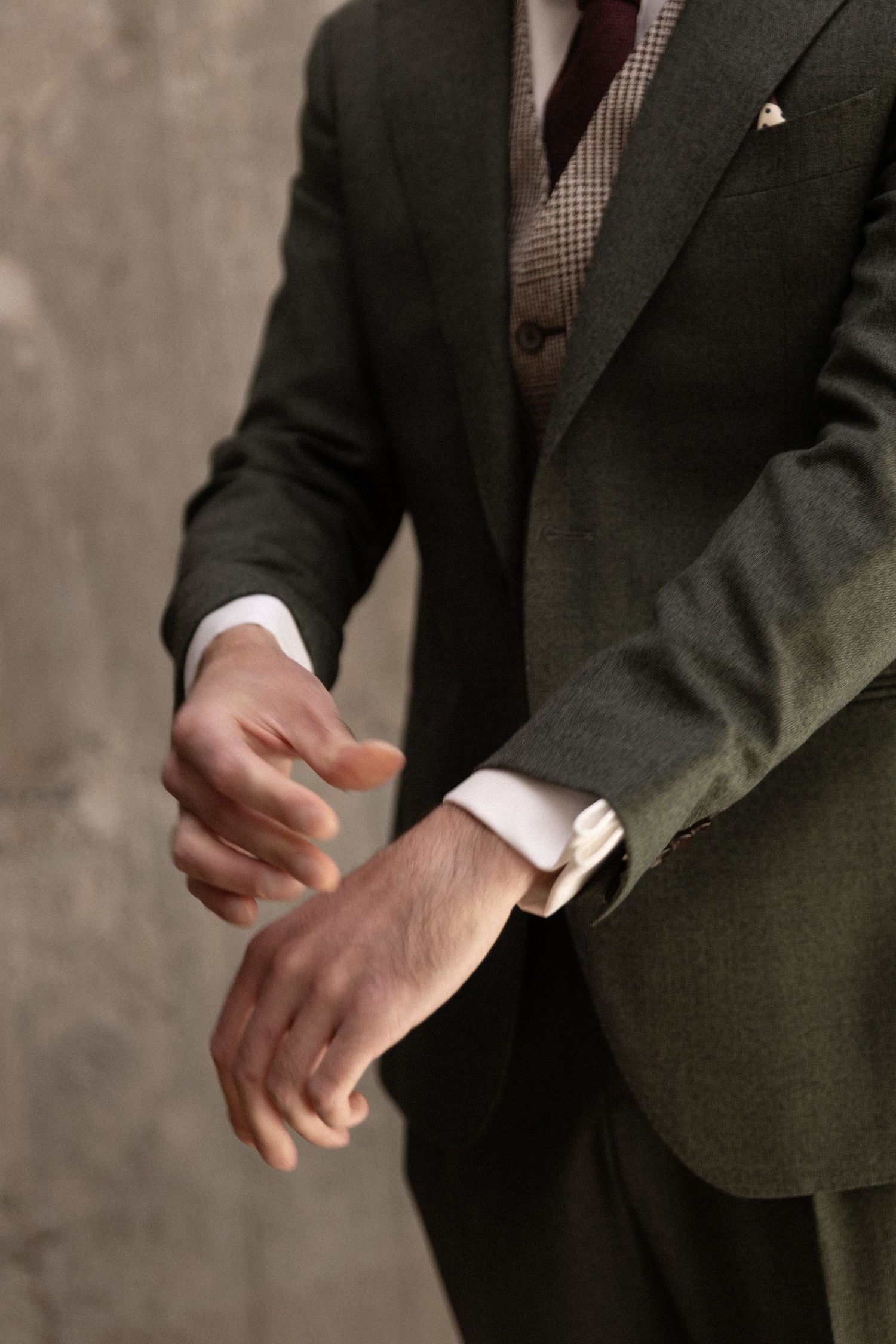 A close-up of hands adjusting a crisp white shirt cuff beneath a dark green suit jacket and brown checkered vest.