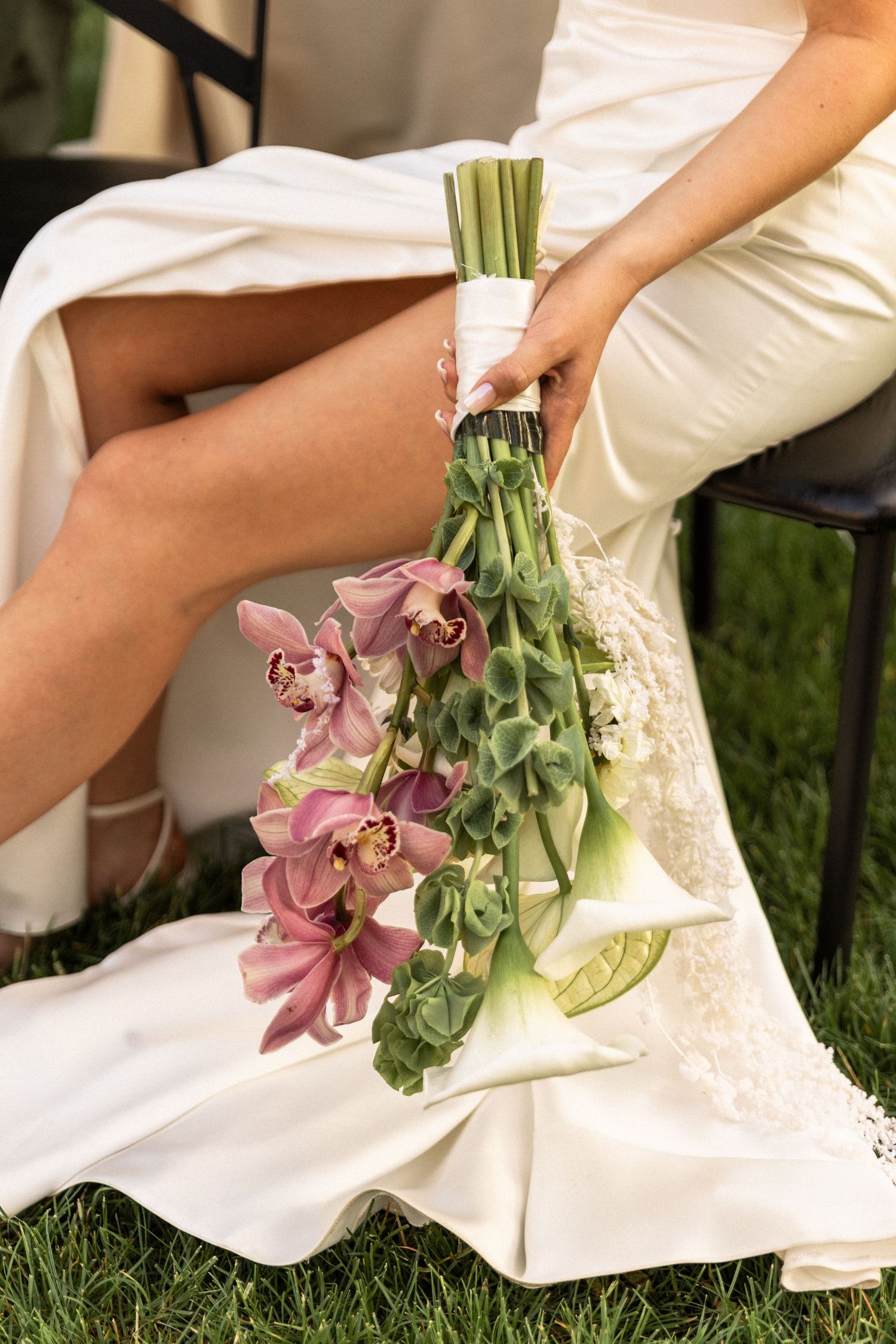 Hand holding elegant bridal bouquet with pink orchids and eucalyptus against white dress and grass background.