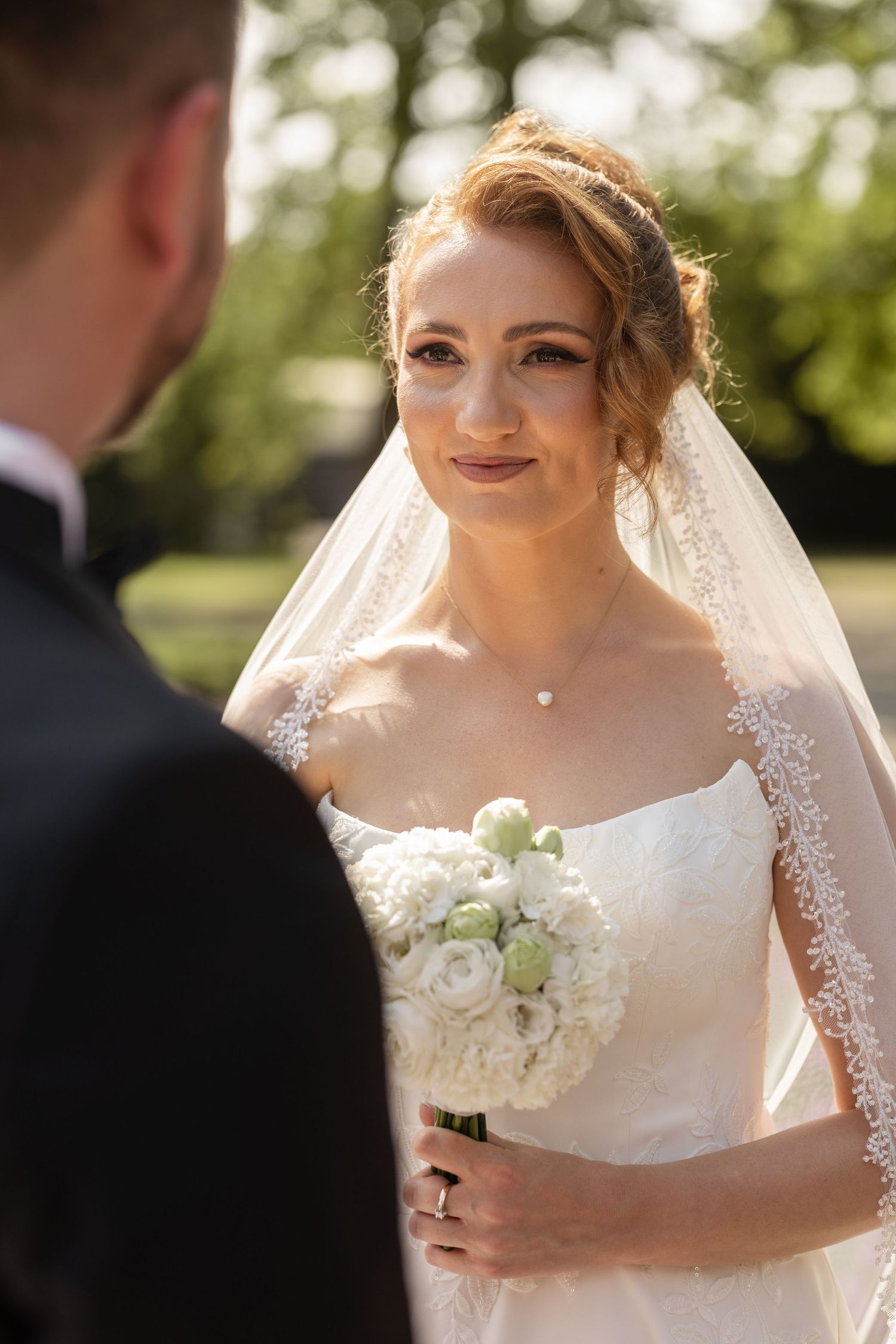 A bride in a white lace wedding dress and veil holds a bouquet of white and cream roses during an outdoor ceremony.