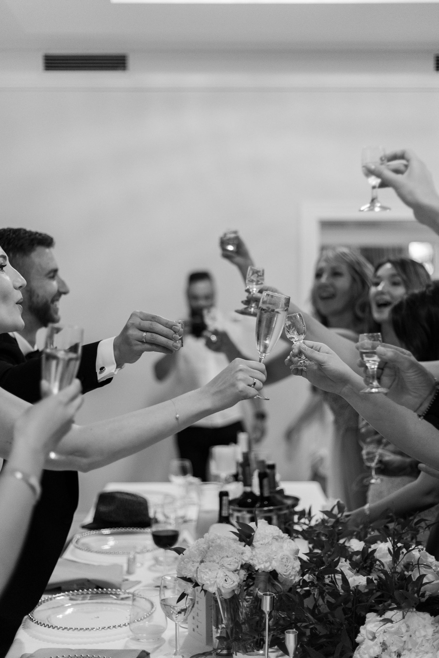 Black and white photo of a celebratory toast at a wedding reception with champagne glasses raised among floral centerpieces.