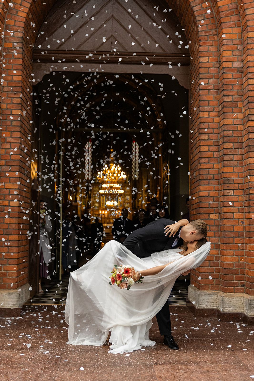 A couple in wedding attire dances beneath brick archways while white flower petals fall around them.