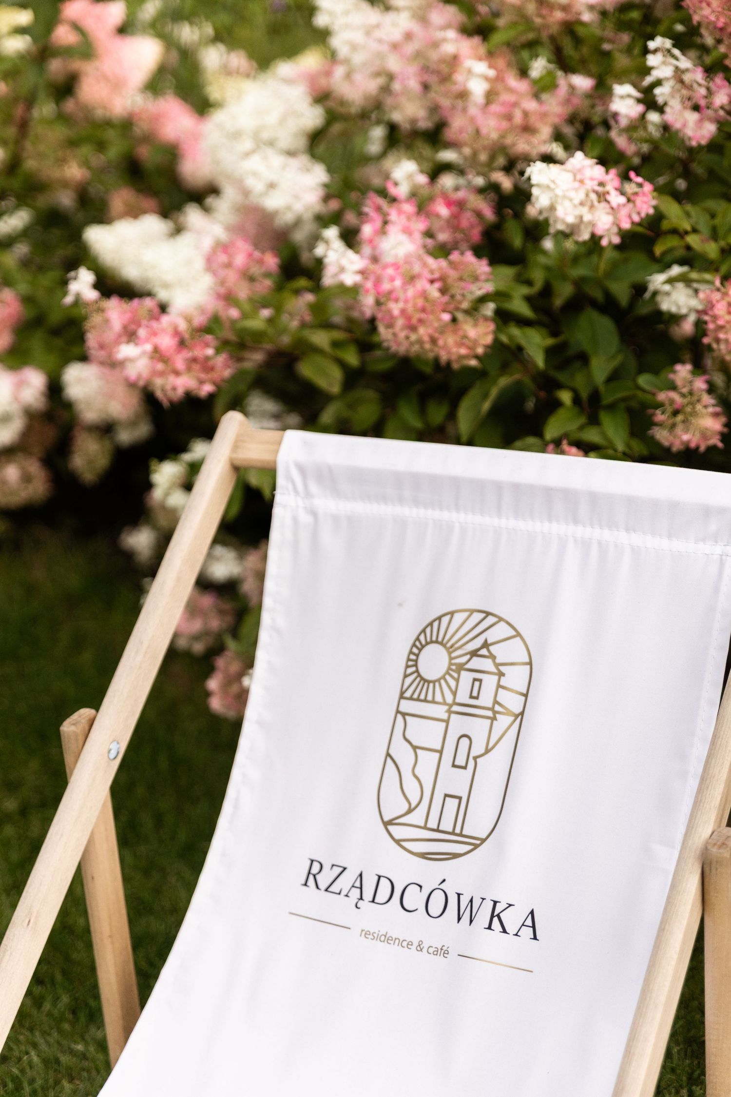 Elegant white deck chairs with gold logo designs arranged under blooming pink cherry blossoms in a garden setting.