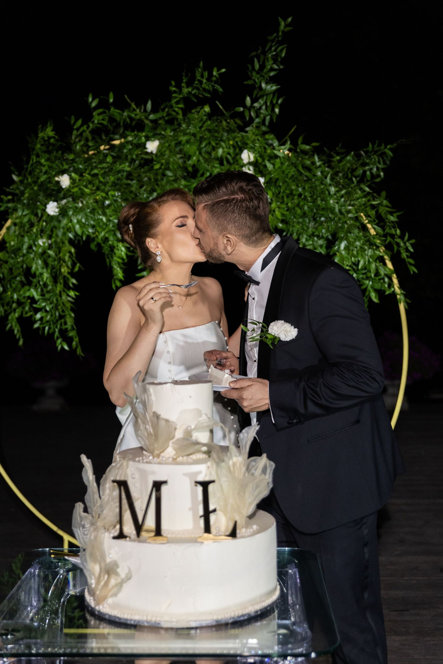 A wedding couple shares a romantic kiss behind their elegant white tiered cake at their evening reception.