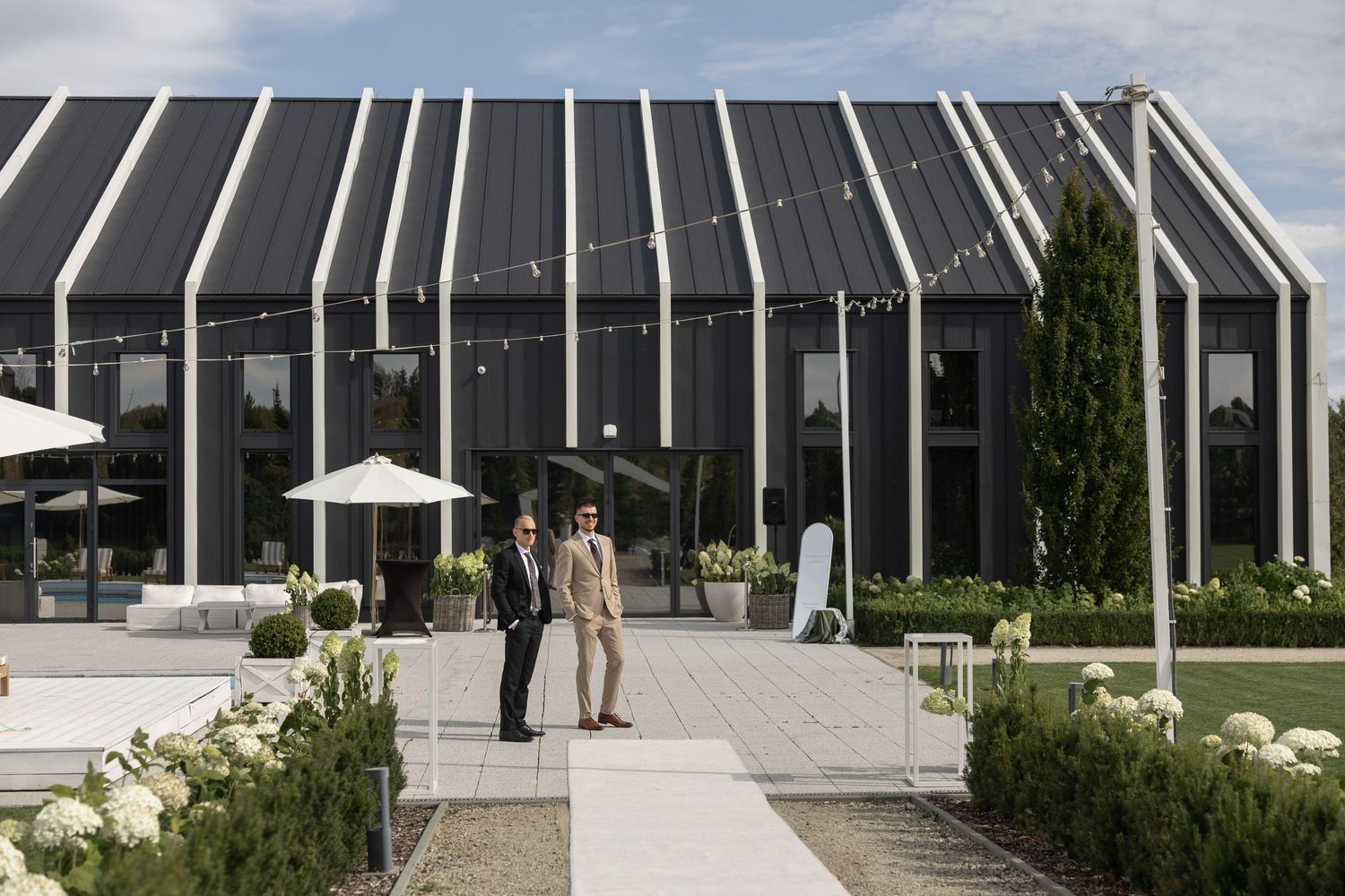 Two people stand outside a modern black and white striped building with string lights and manicured gardens.
