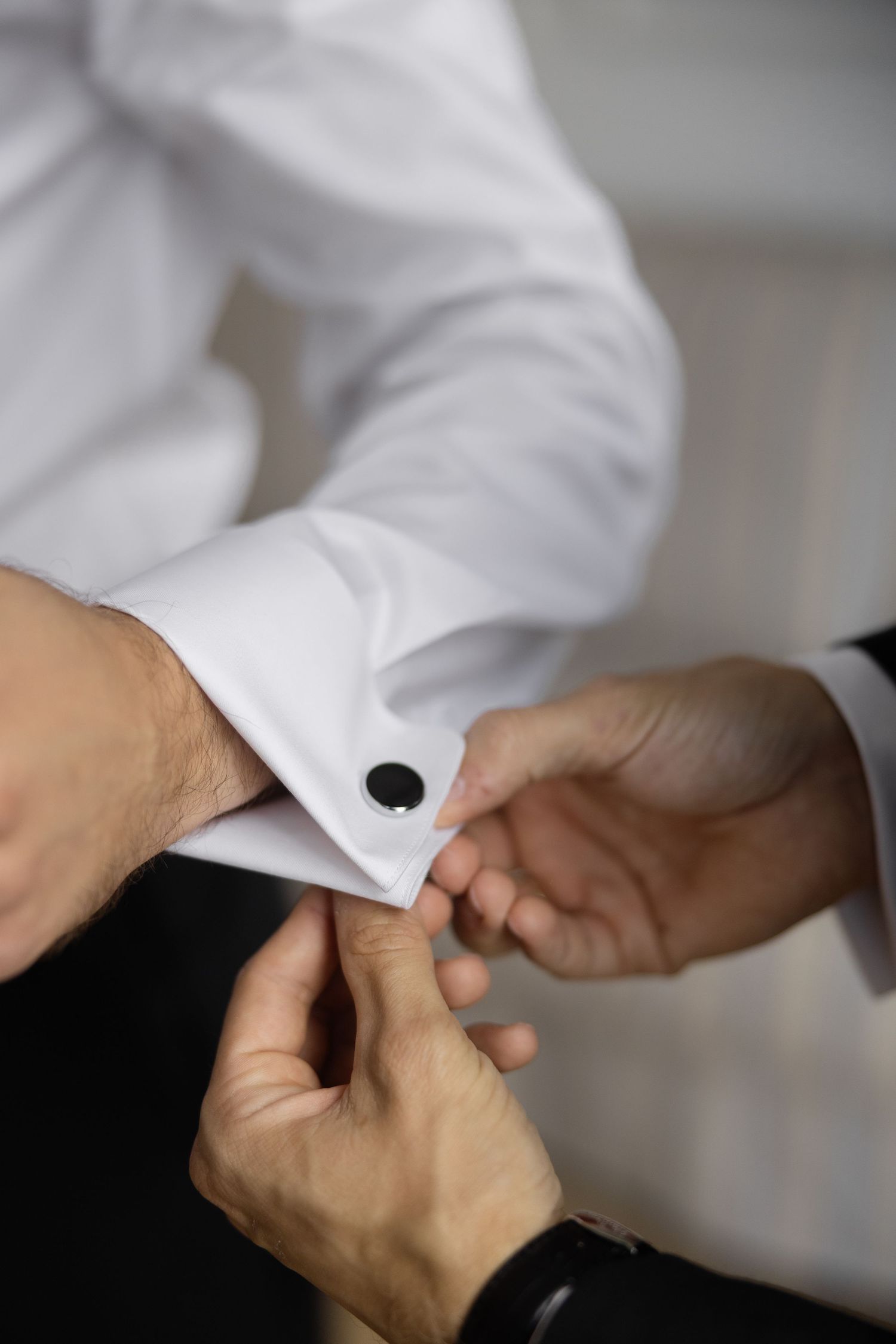 A person fastening a black cufflink on a white dress shirt sleeve, assisted by another person's hands.