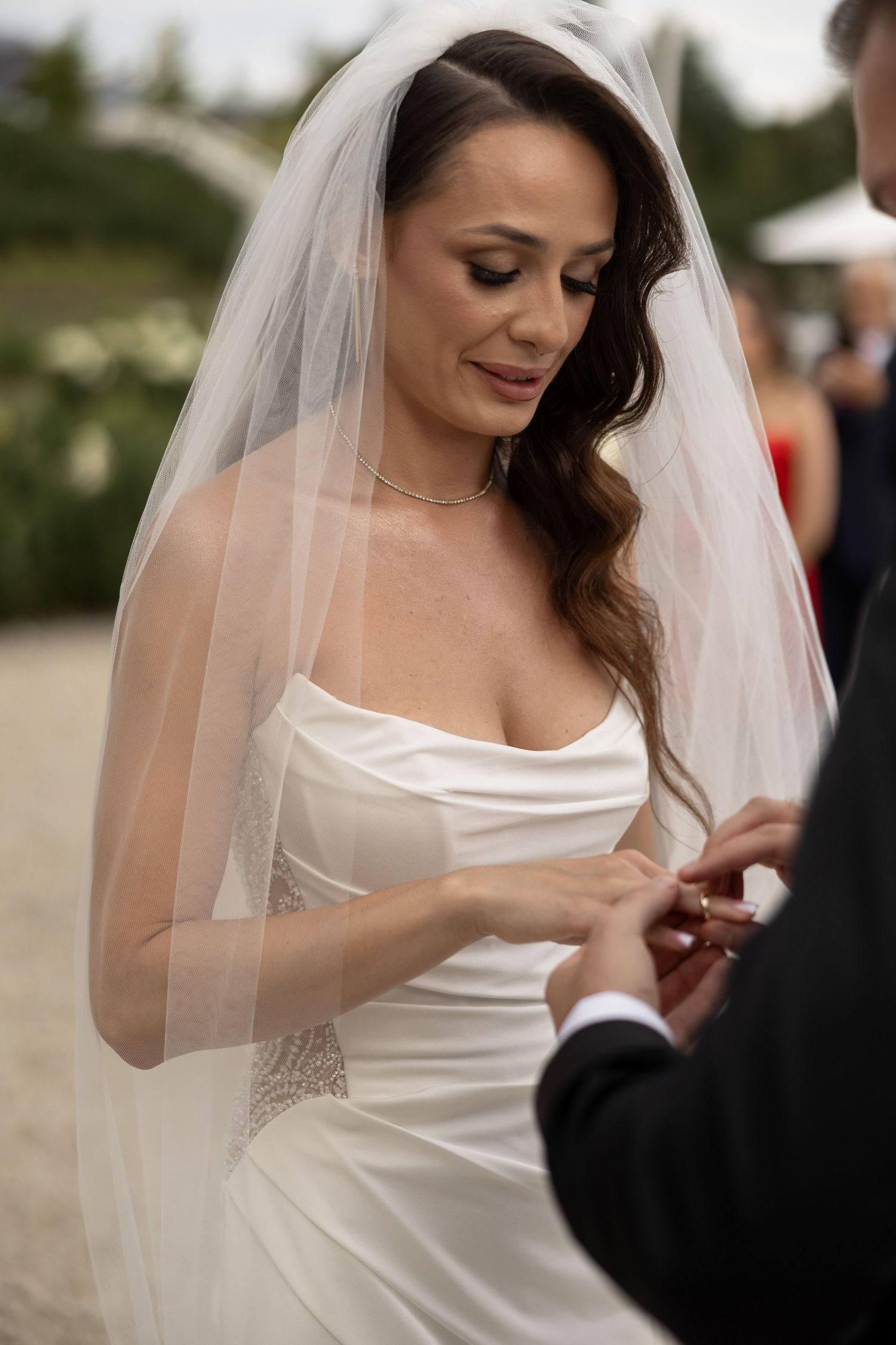 Bride in white satin gown and veil placing wedding ring on groom's finger during outdoor ceremony.