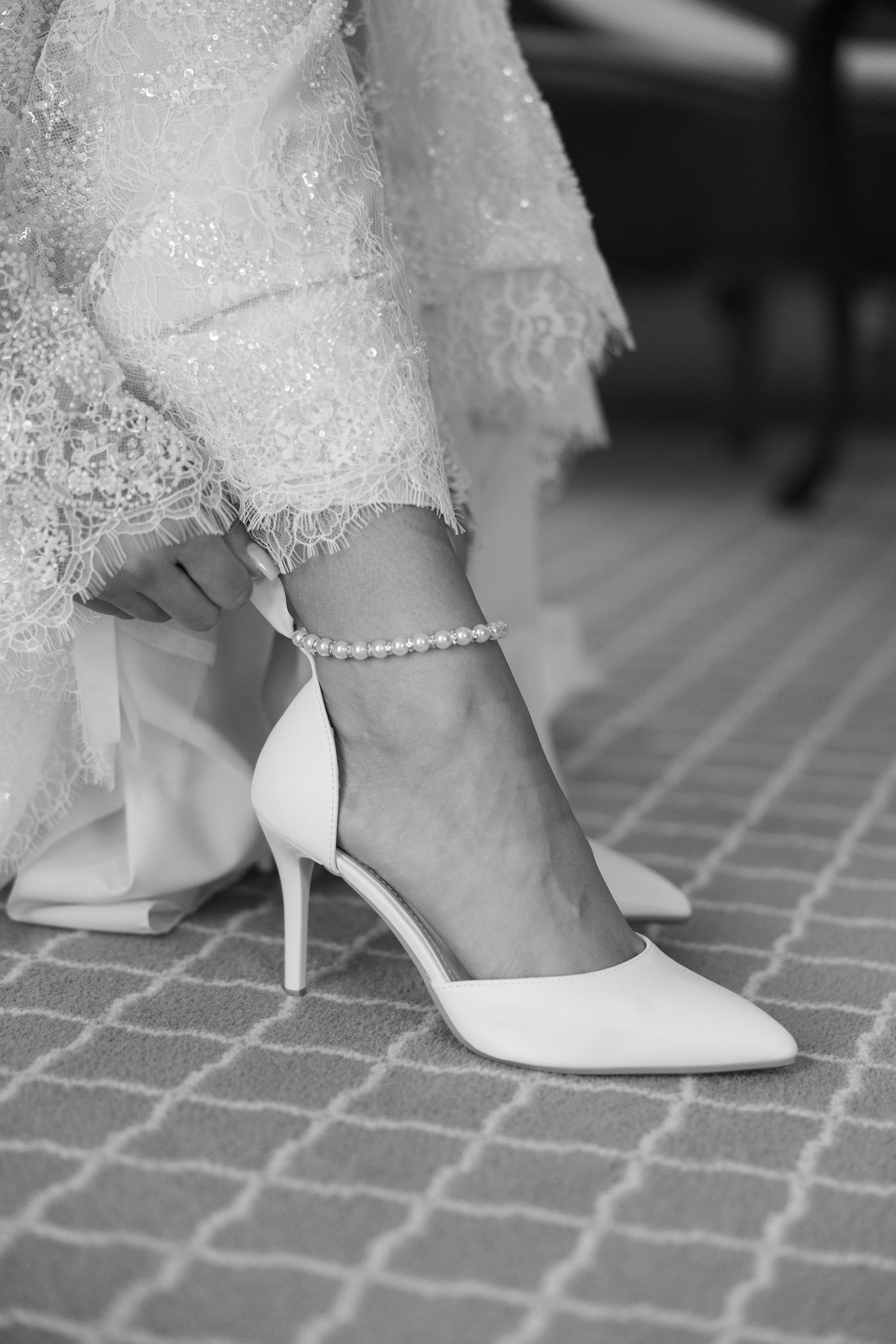 Black and white photo of a bride wearing white pointed-toe heels and a pearl anklet with lace wedding dress.