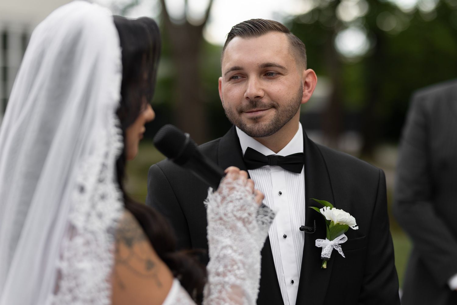 Groom in black tuxedo smiling at bride during outdoor wedding ceremony with white floral boutonniere.