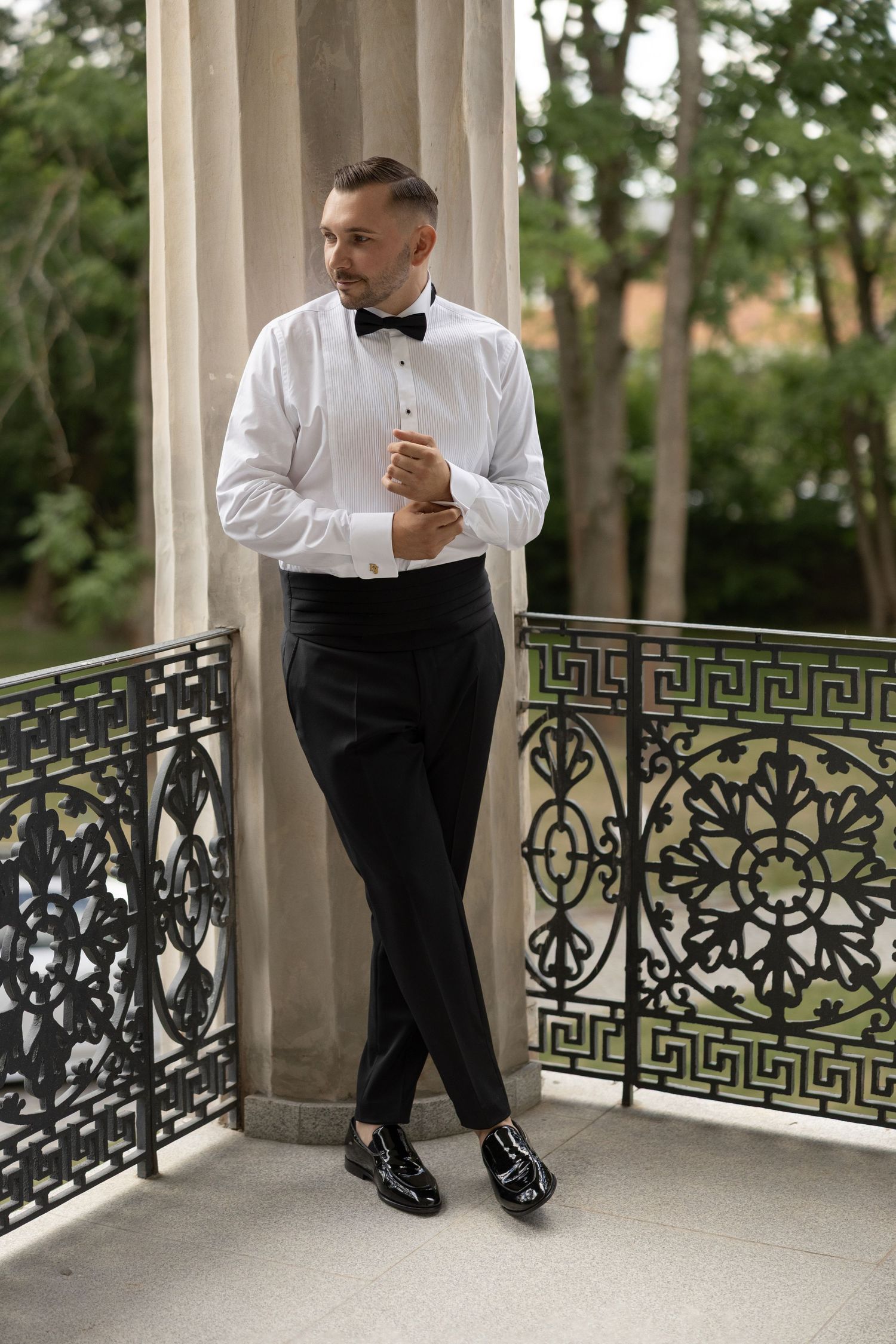 Elegant man in tuxedo shirt, bow tie, and black trousers standing on ornate balcony adjusting cufflinks.