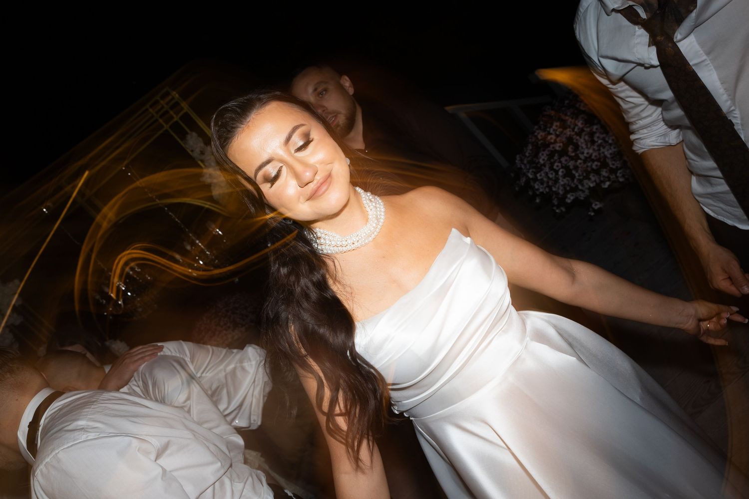 Bride in white gown dancing joyfully at wedding reception, hair flowing, eyes closed, smiling blissfully.