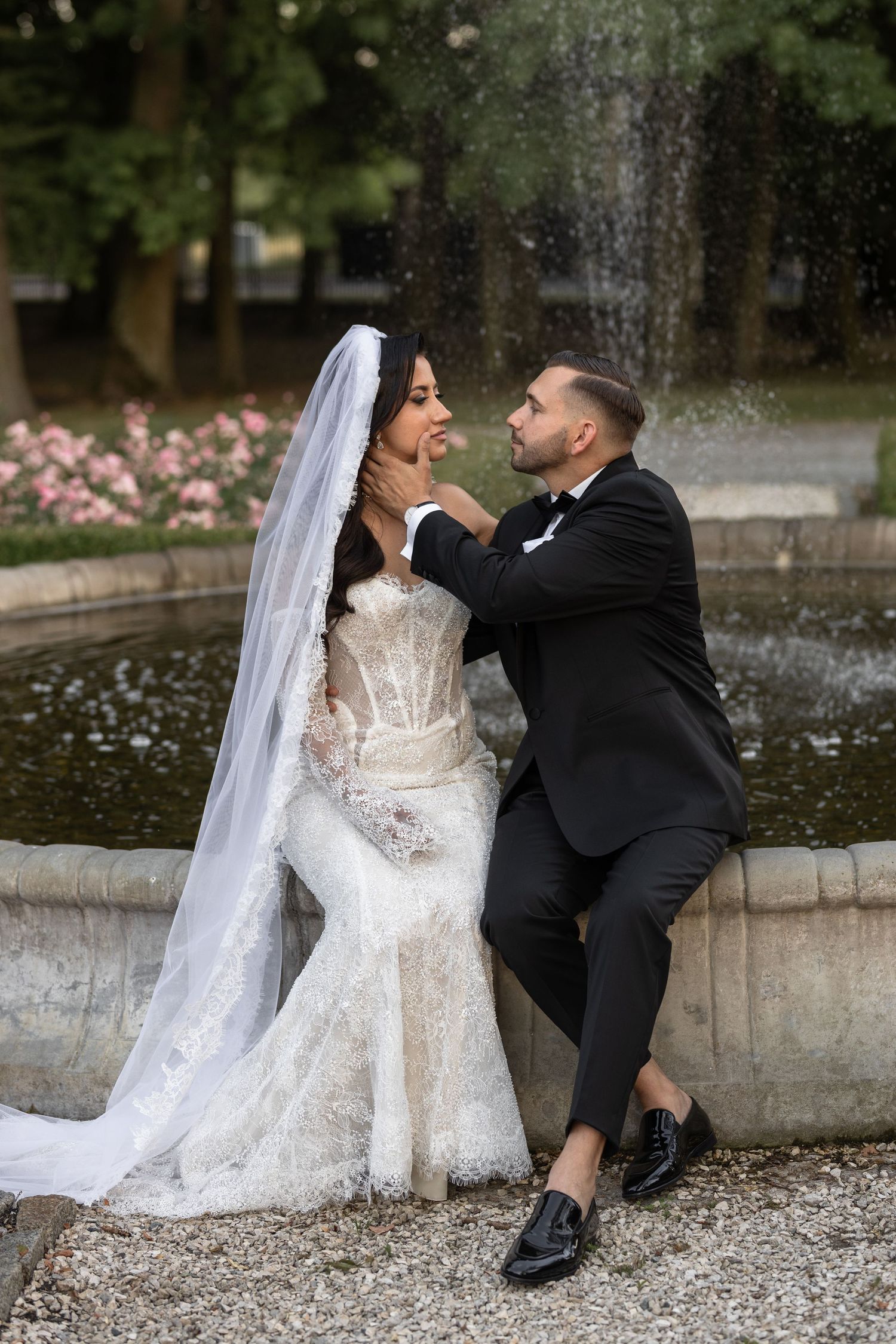 Bride in lace gown and veil with groom in black tuxedo sitting by fountain in garden wedding photo.