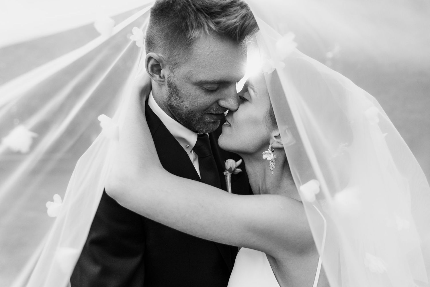 Black and white romantic photo of newlyweds sharing a tender kiss under a flowing veil.