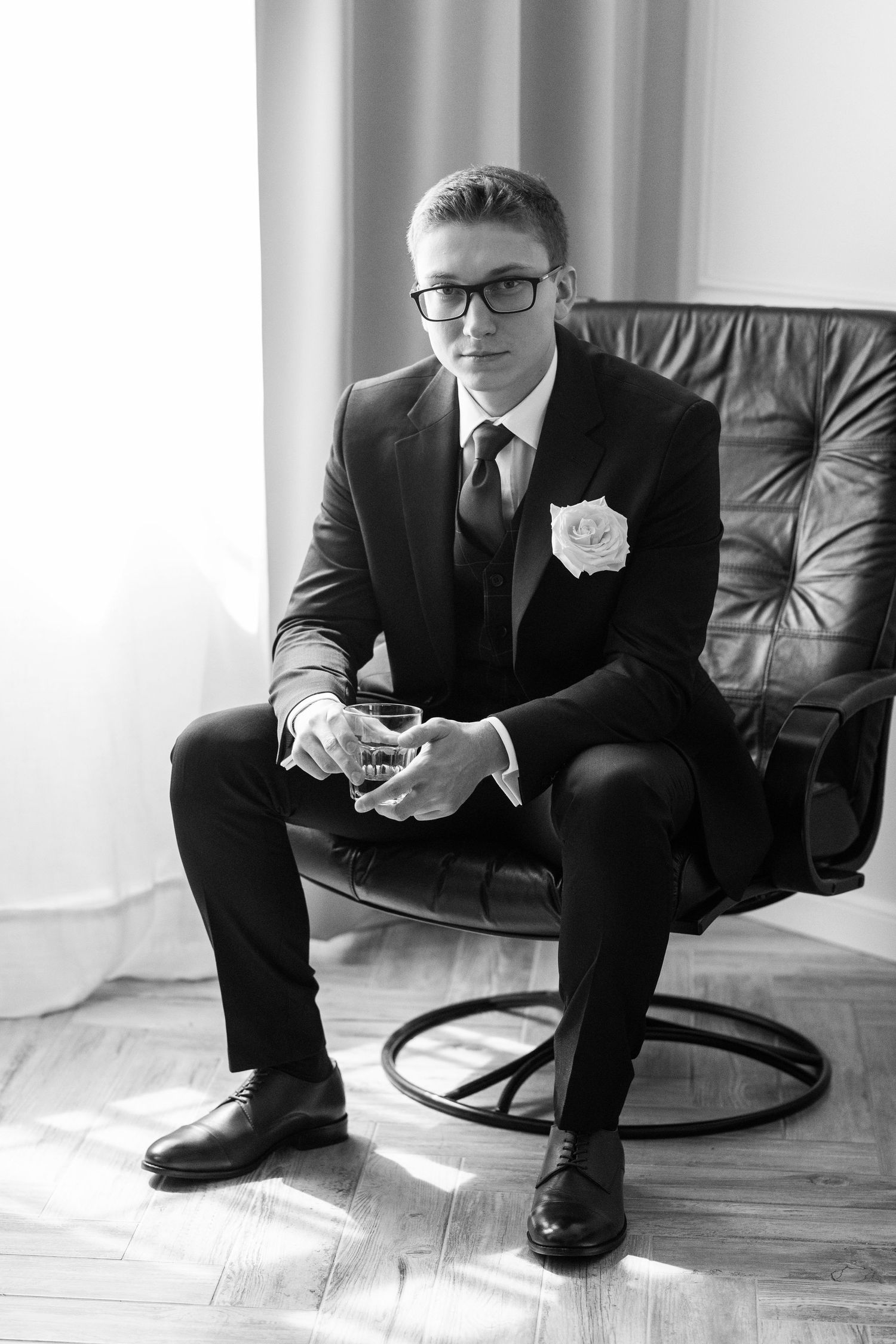 Black and white portrait of someone in a formal suit and tie sitting on a leather chair with a boutonniere.