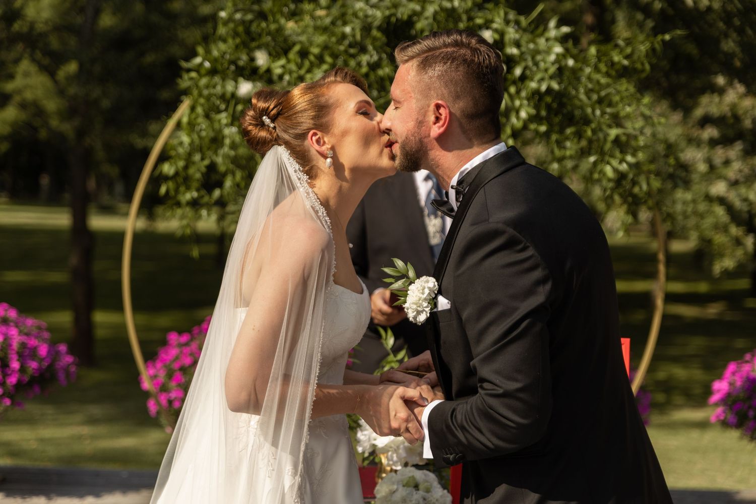 A romantic wedding kiss in a garden setting with pink flowers and a decorative arch in the background.