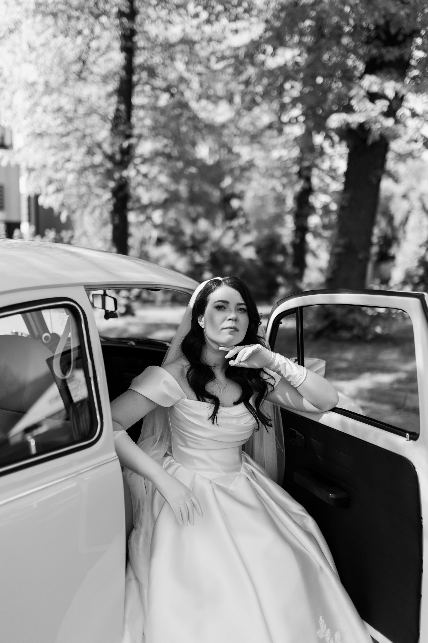 A bride in an elegant white off-shoulder gown poses gracefully inside a vintage car on her wedding day.