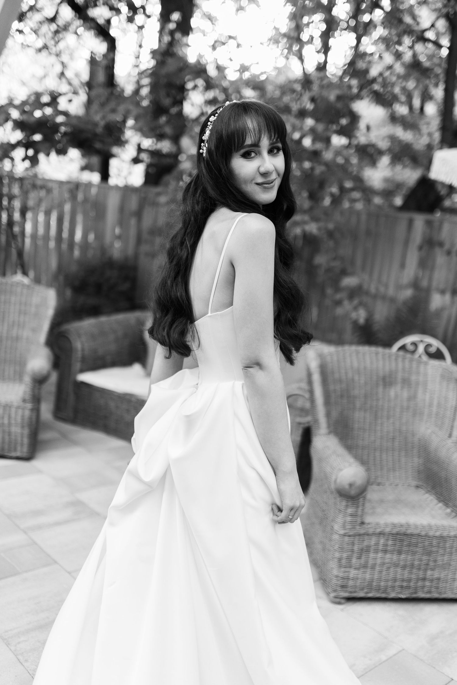 Black and white portrait of someone in an elegant white wedding dress with long dark hair looking over their shoulder on a patio.