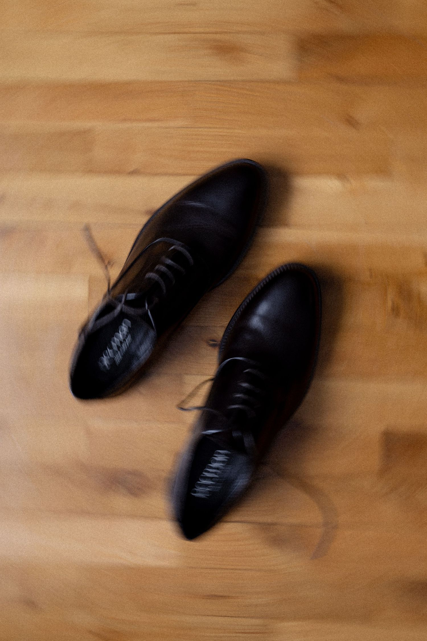A pair of black leather dress shoes with laces on a light wooden floor.
