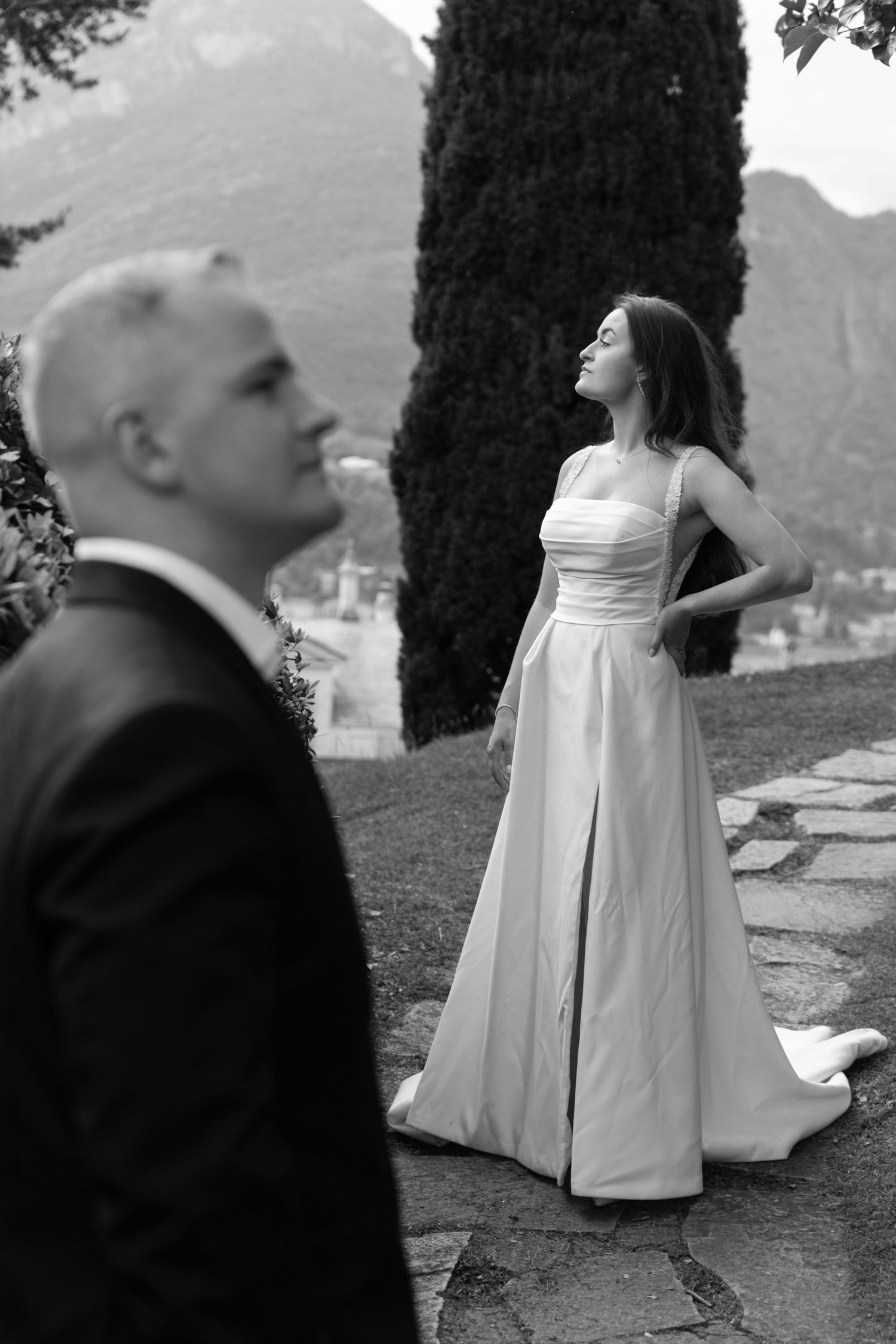 Black and white photo of bride in elegant gown posing outdoors with groom in foreground, mountains and cypress tree behind.