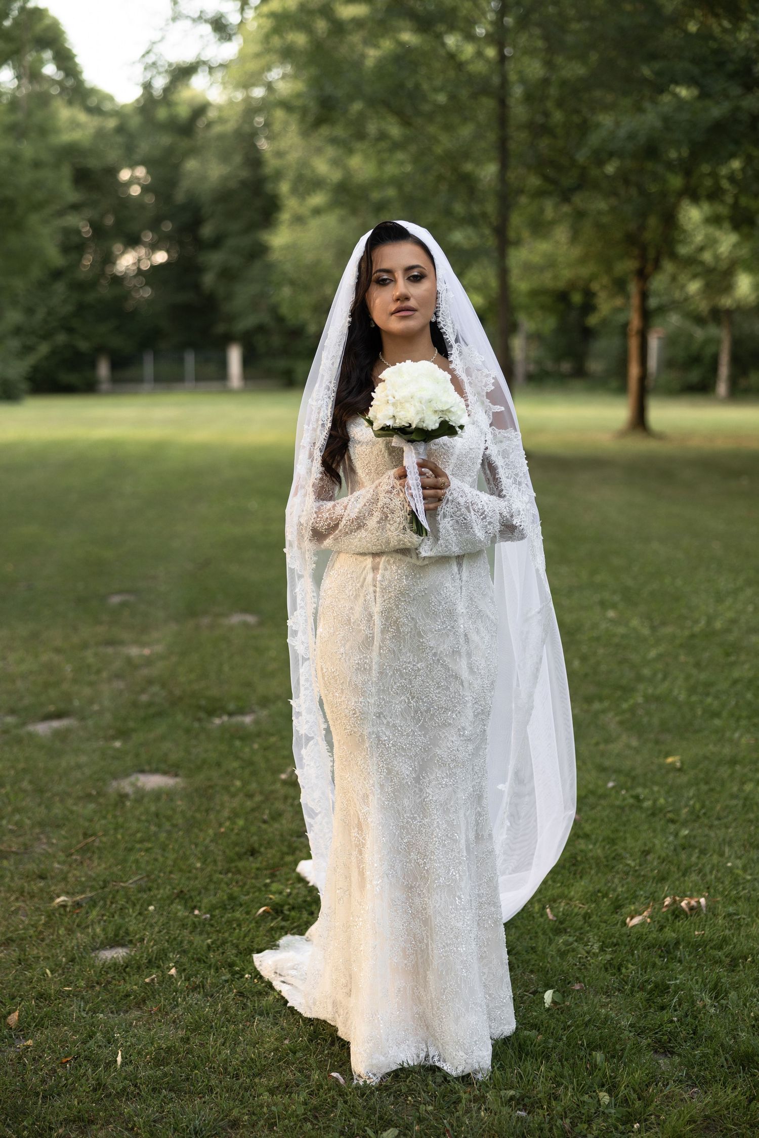 Bride in lace wedding gown and veil holding white bouquet standing on green grass in park.