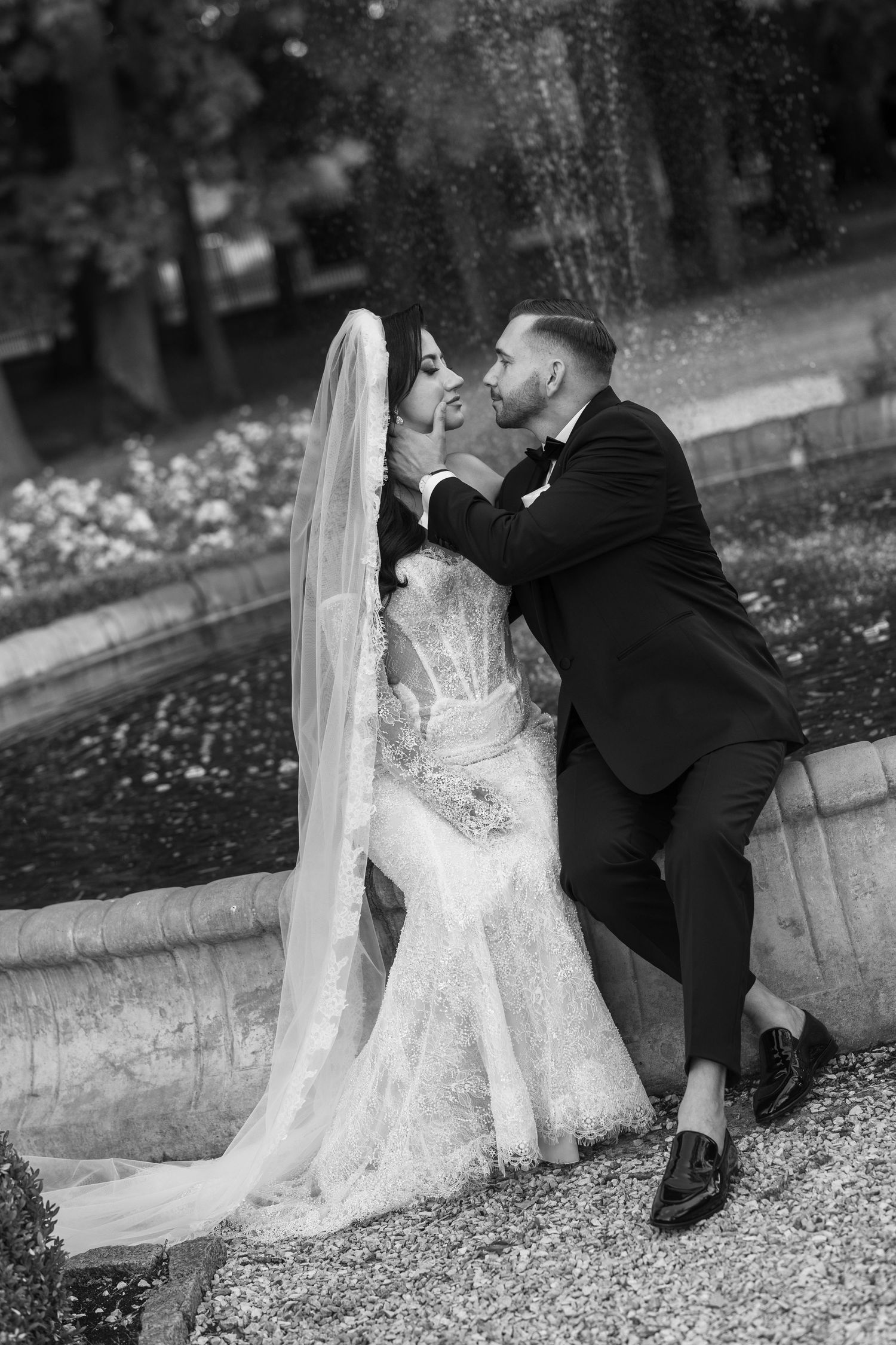 Black and white photo of a bride and groom sharing a romantic moment by a fountain, bride in lace gown with veil.