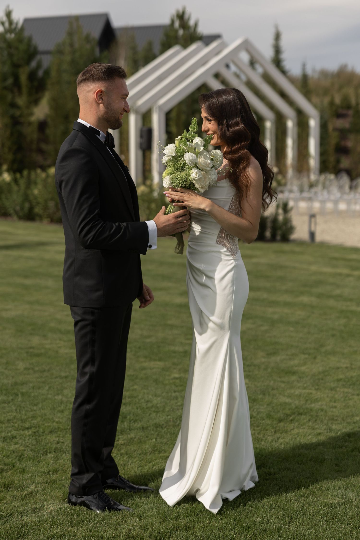 Bride in white gown holds bouquet while groom in black tuxedo stands facing her on green lawn at outdoor wedding.