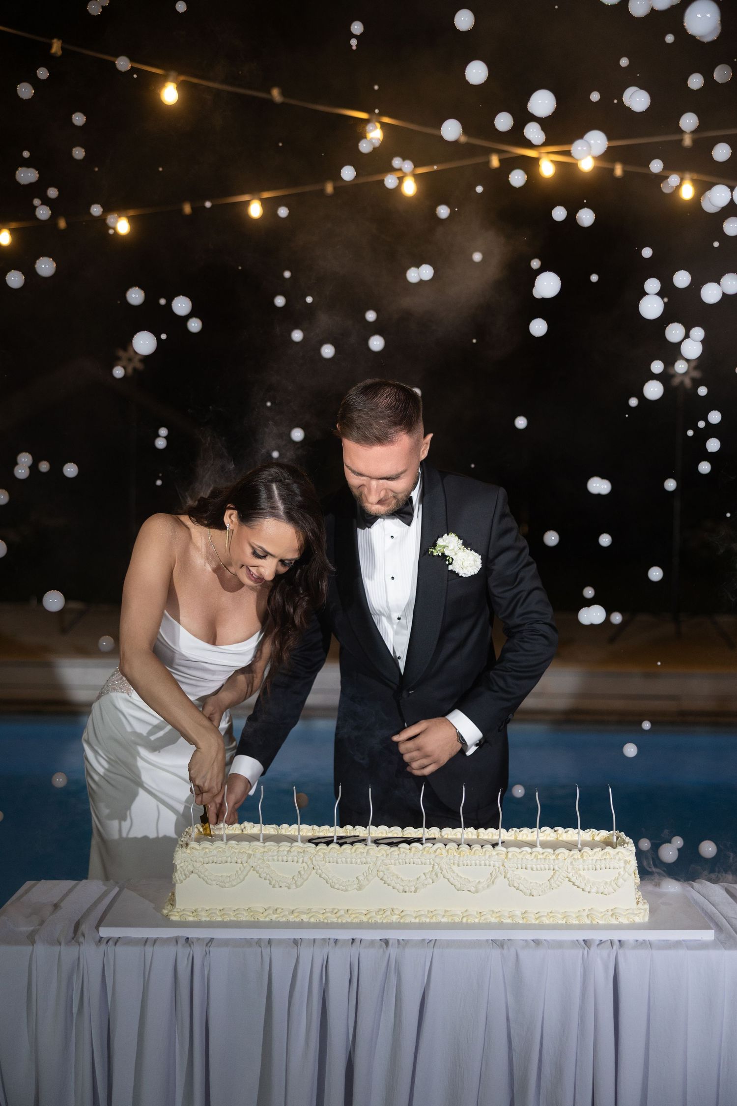 Bride and groom cutting wedding cake at night with bokeh lights in background.