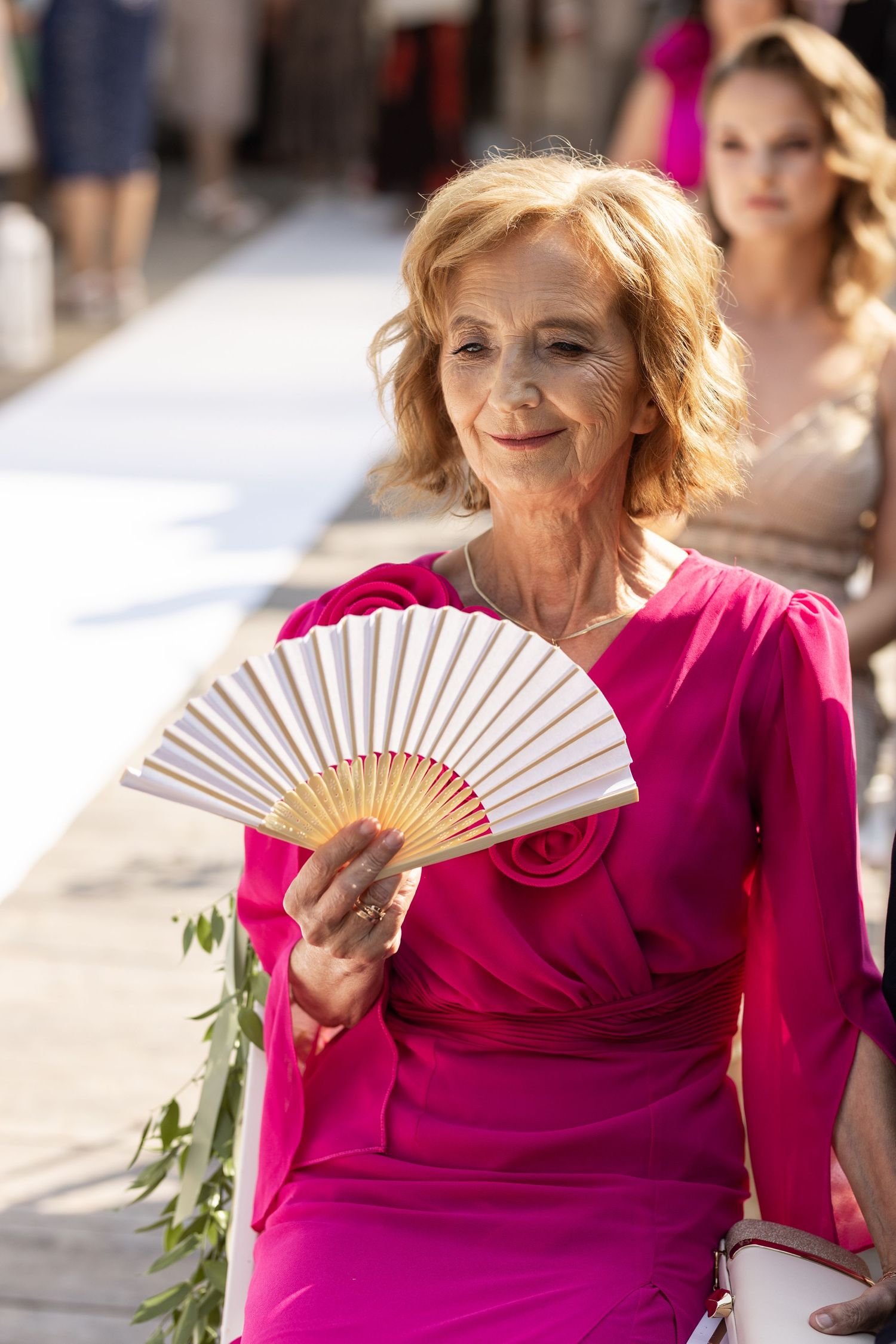 Woman in vibrant pink dress poses elegantly with decorative white hand fan in outdoor setting.