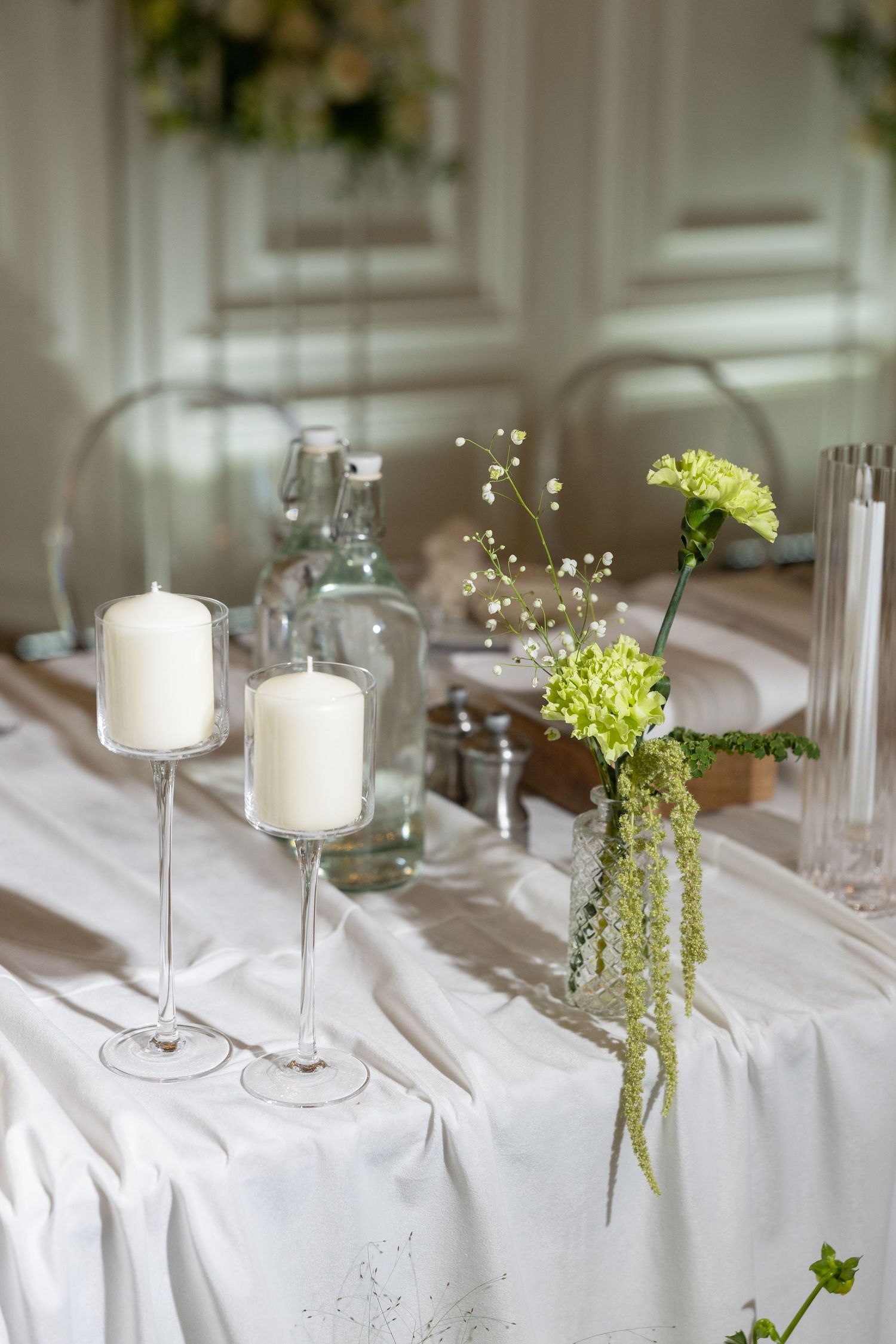 Elegant wedding table with white candles on glass holders, green floral arrangement, and white linen tablecloth in a bright venue.