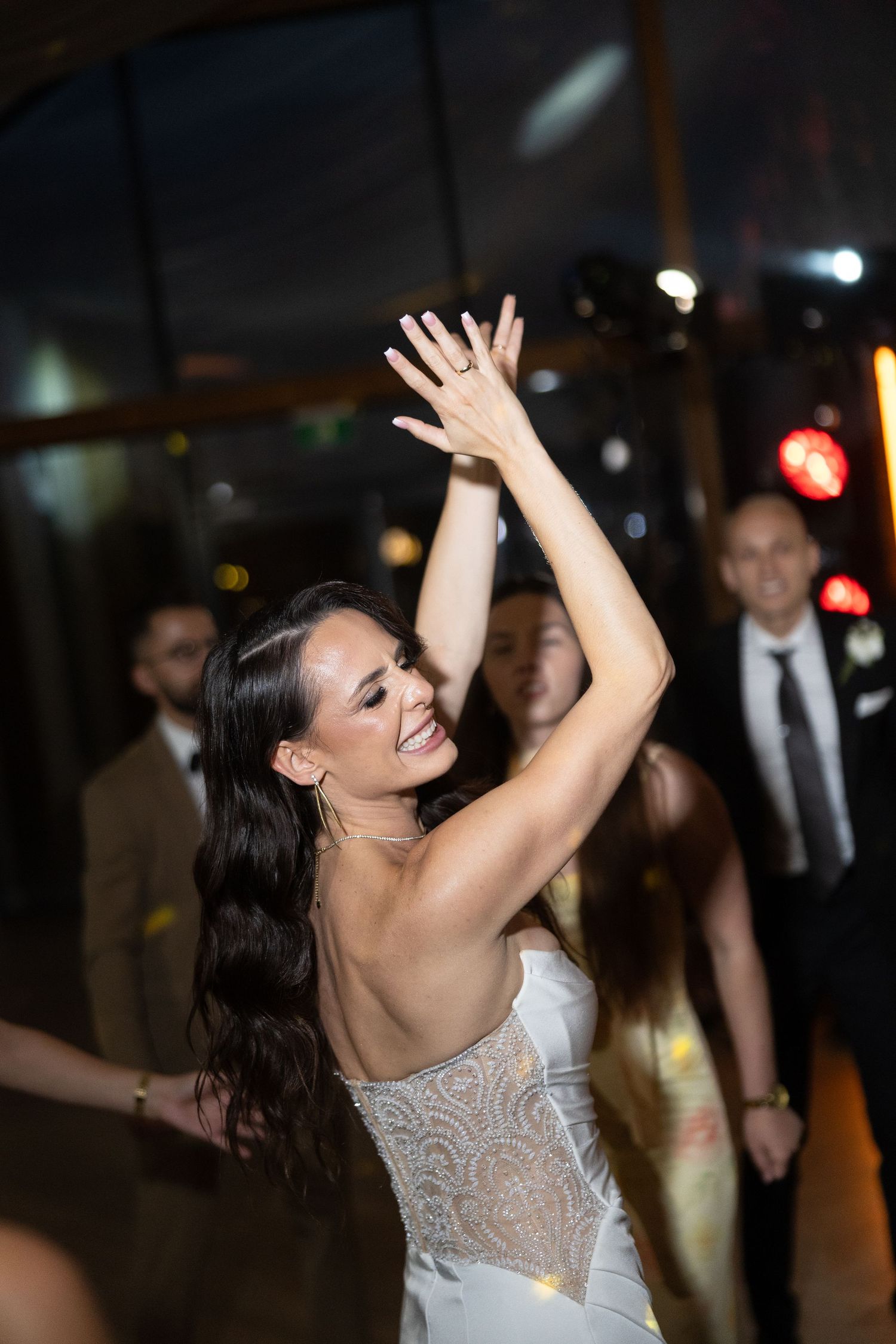 Bride in a backless lace wedding gown dancing joyfully with arms raised at a wedding reception.