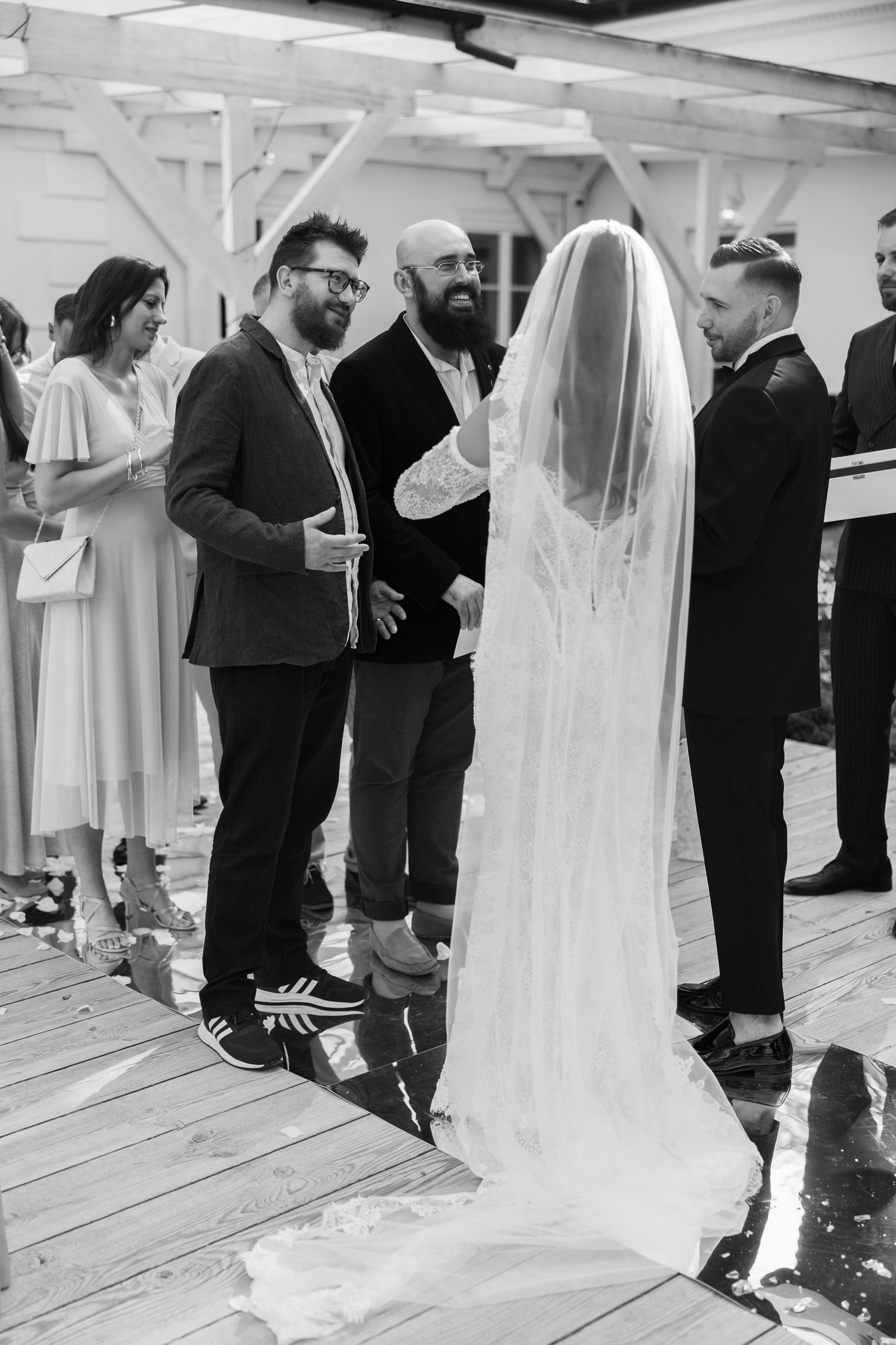Black and white wedding ceremony photo showing bride in veil with groom and guests on wooden deck.