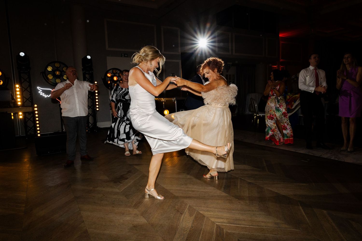 Two wedding guests dance joyfully on a dark wooden dance floor as a photographer captures the moment with flash.