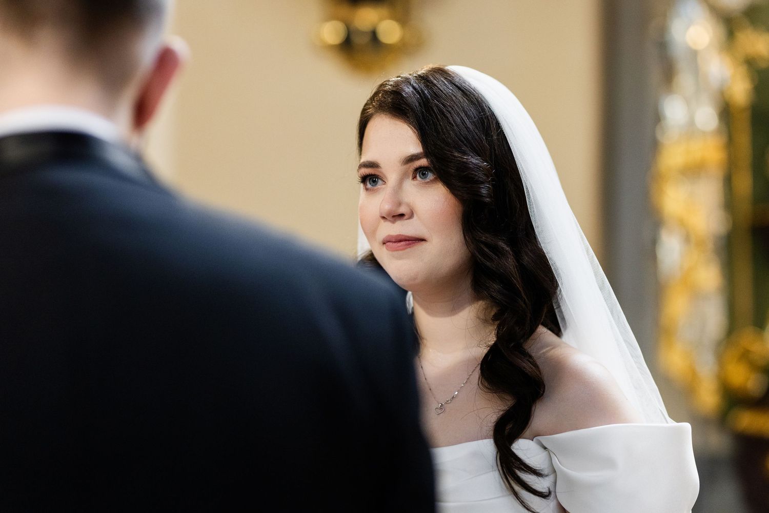 A bride in a white wedding dress with wavy dark hair and veil gazes lovingly at her groom during their wedding ceremony.