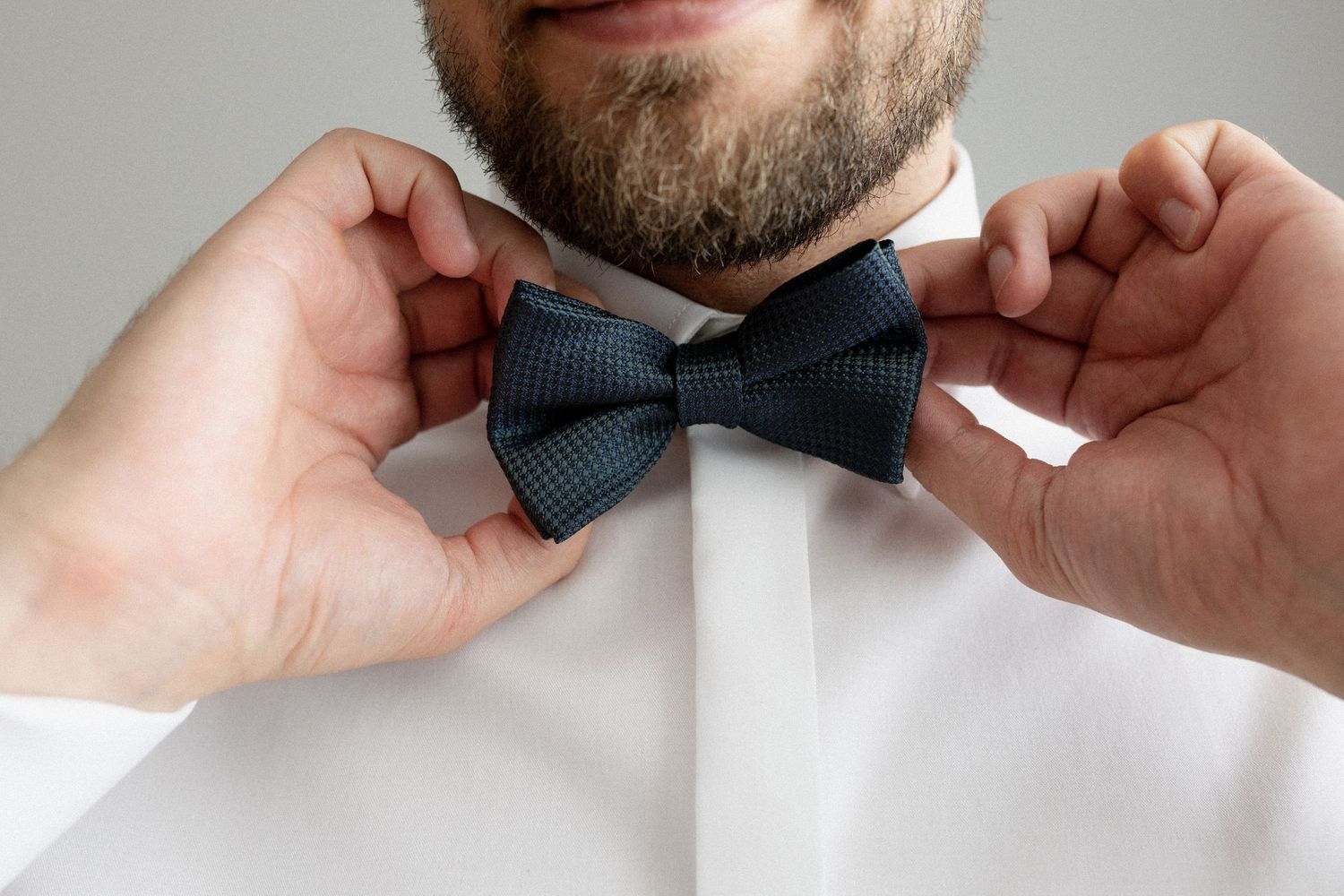 Hands adjust a dark blue bow tie against a crisp white dress shirt in a formal attire preparation shot.