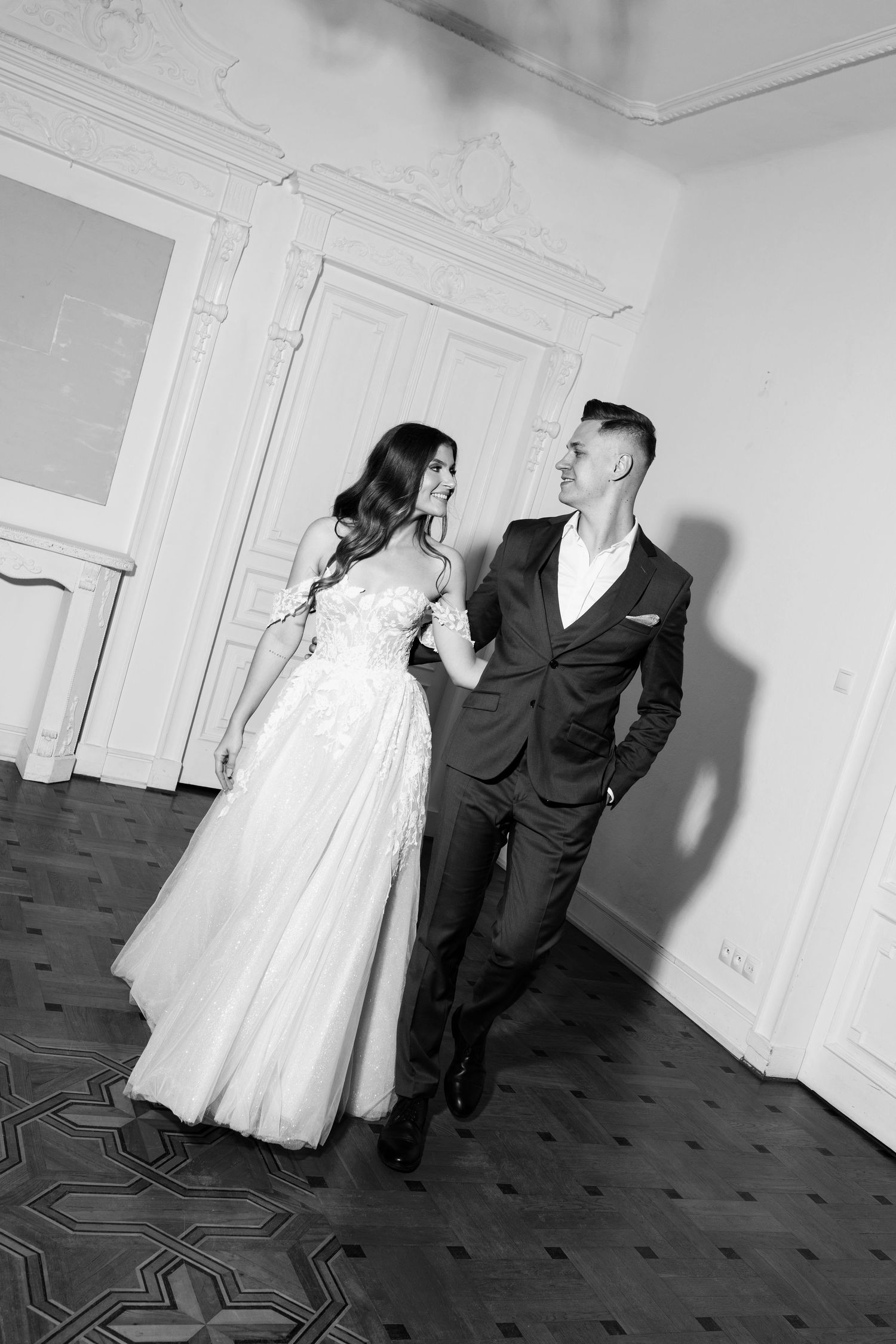 Black and white photo of elegantly dressed couple in formal attire standing in ornate room.