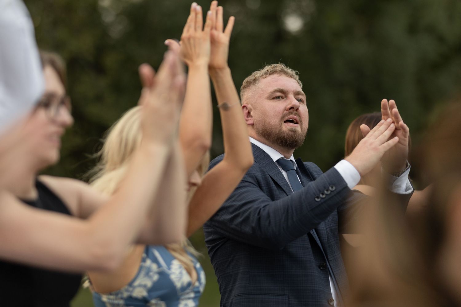 Man in navy suit clapping and singing outdoors at a celebration with guests raising hands around him.