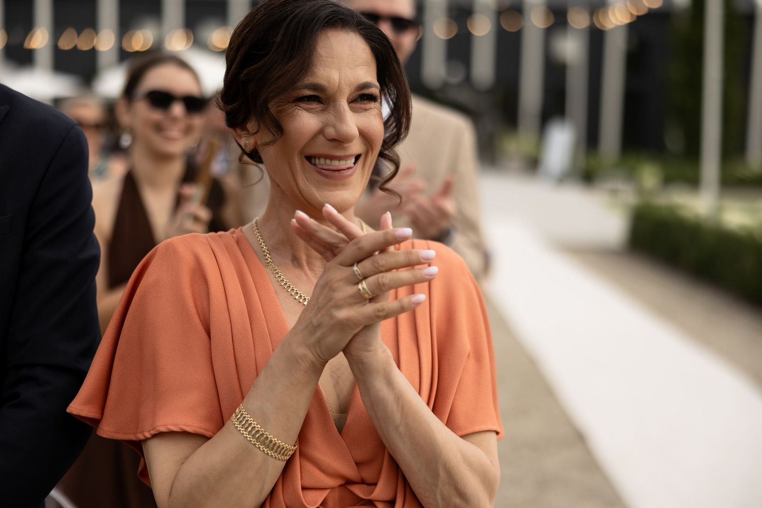 Woman in orange dress clapping and smiling at outdoor event, wearing gold jewelry and rings.