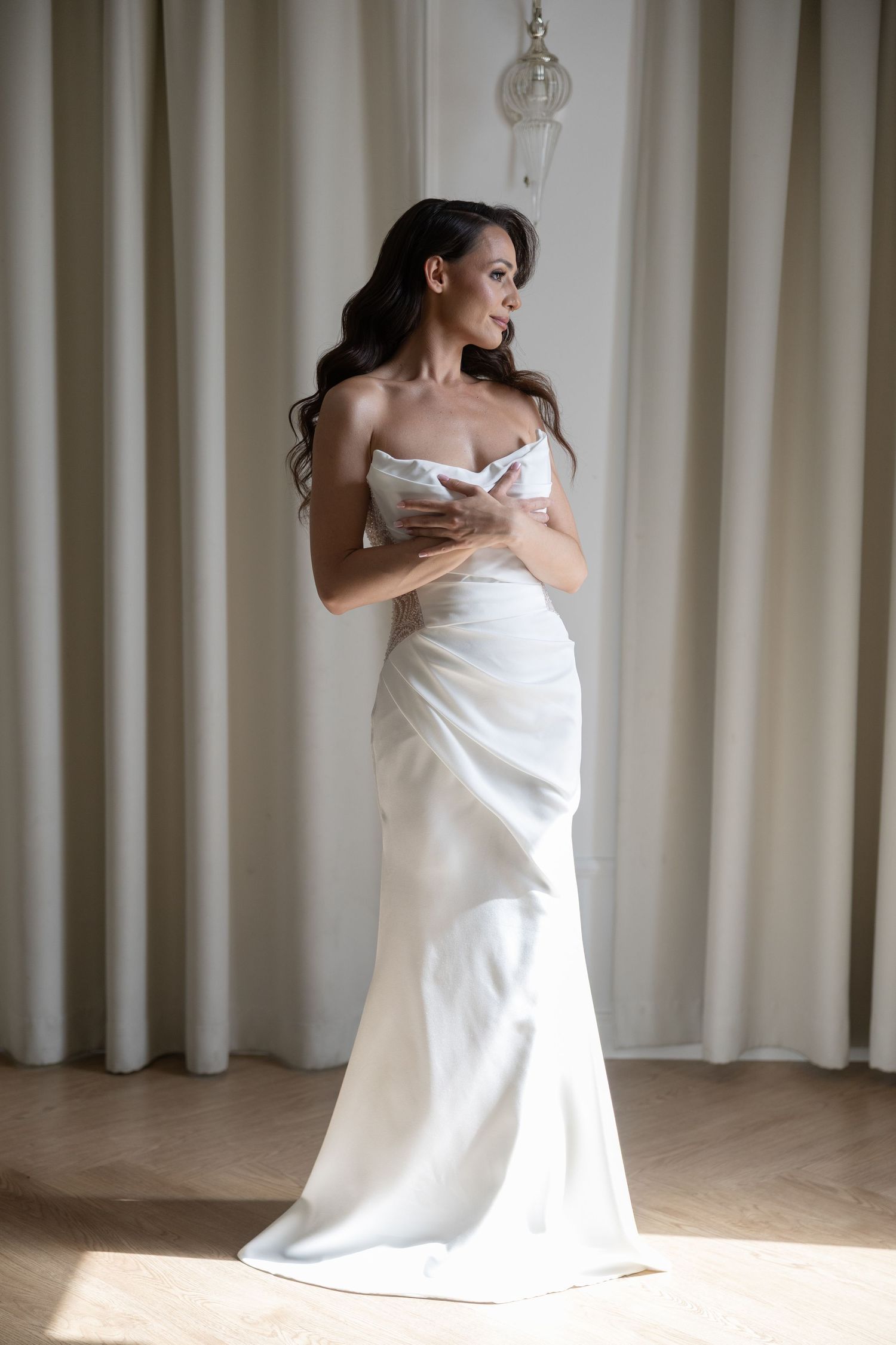 Elegant bride in a strapless white satin mermaid wedding gown standing gracefully before sheer curtains.
