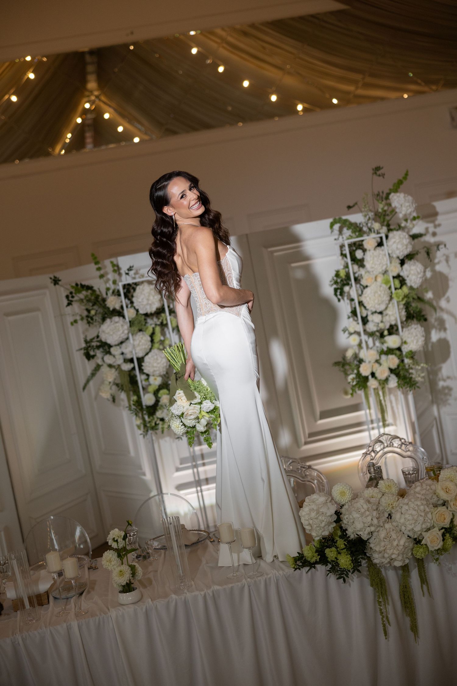 Elegant bride in white gown holding bouquet stands among white floral arrangements in a beautifully decorated wedding venue.