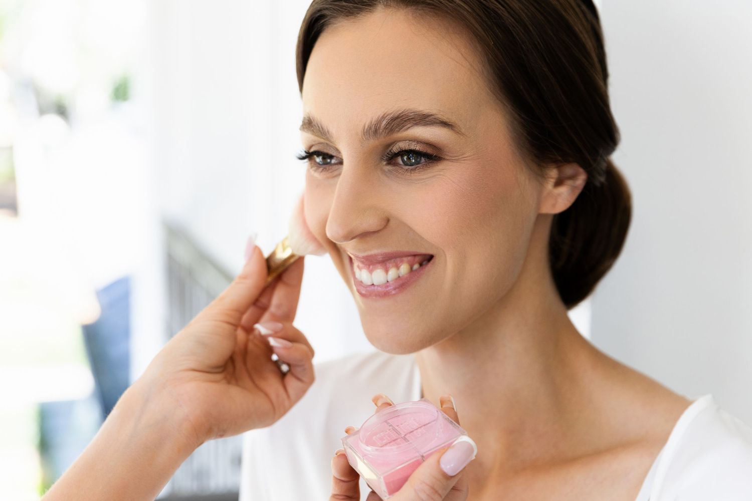 Elegant woman in white top smiles while gently touching her face and holding a pink skincare product.