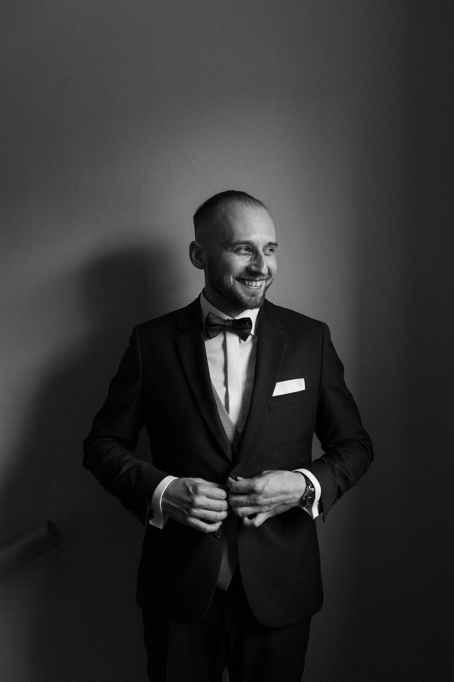 A black and white portrait of someone in a classic tuxedo with bowtie smiling to the side against a dark background.