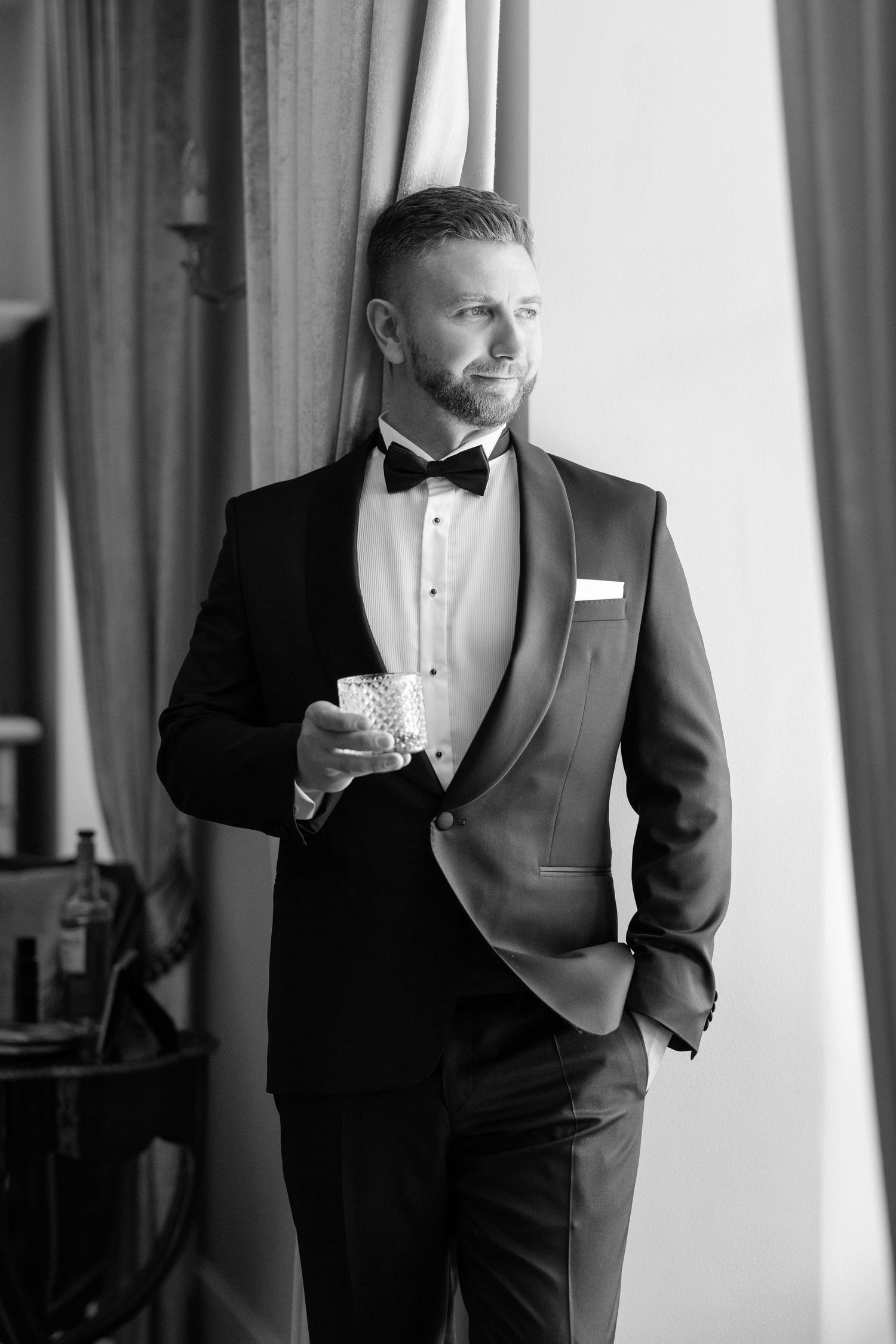 A groom in a classic black tuxedo poses thoughtfully by a window in an elegant black and white photograph.