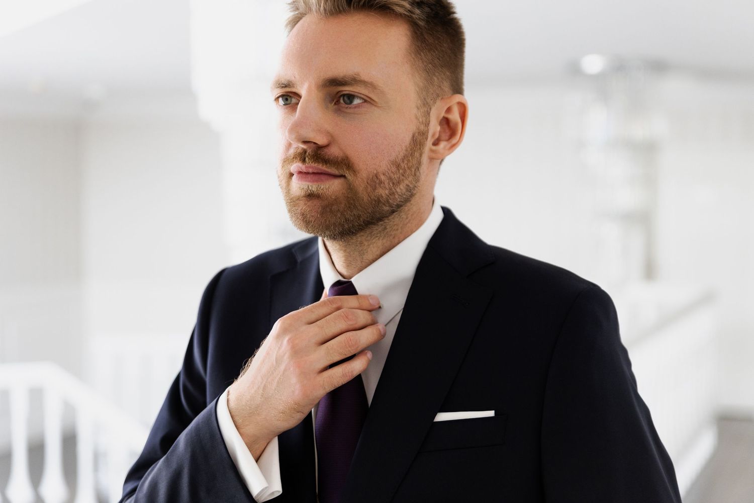 A well-dressed person in a black suit adjusts their burgundy tie while standing in a bright modern interior.