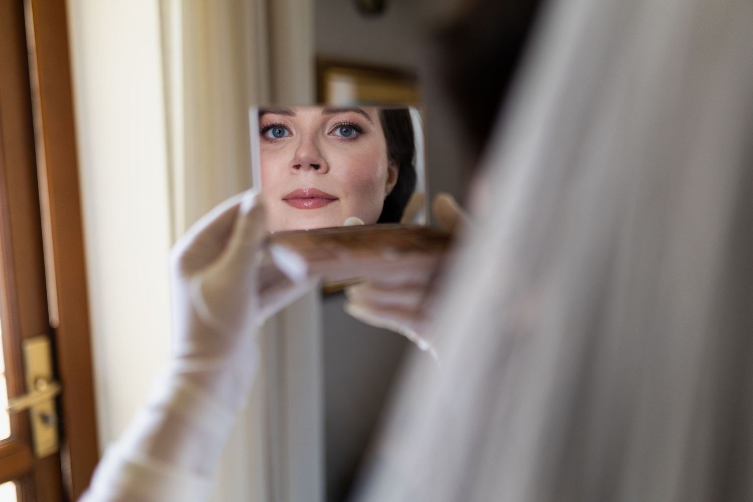A small handheld mirror reflects someone in soft natural light looking at their reflection through delicate white fabric.