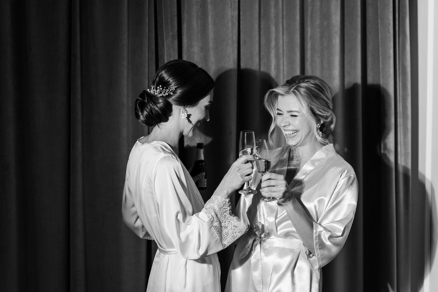 Two women in silky white robes laugh and clink champagne glasses together in black and white photograph.