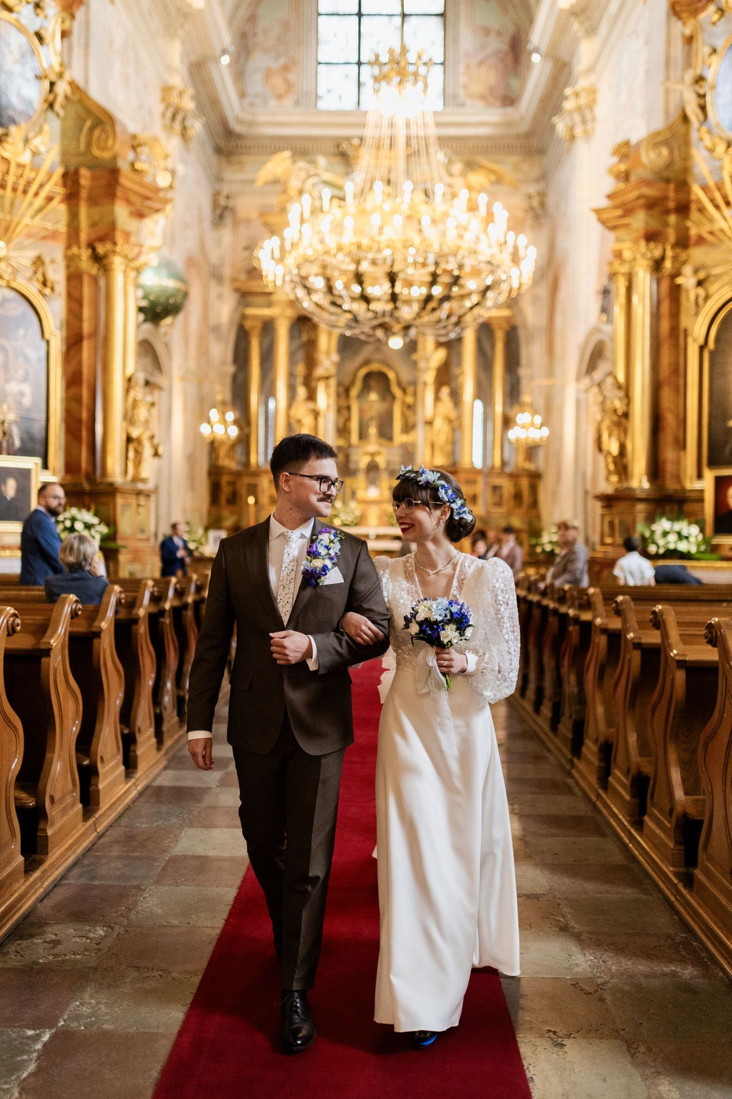 A couple walks down the aisle of an ornate baroque church with crystal chandeliers and gilded decorations.