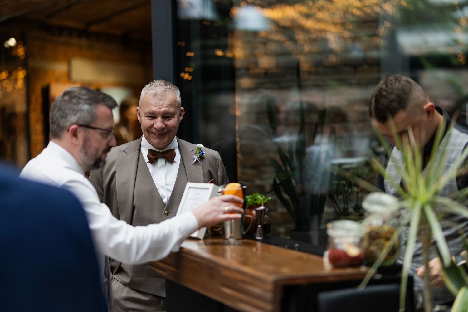 Two men share drinks and conversation while seated at a wooden bar counter near glass windows in a restaurant.