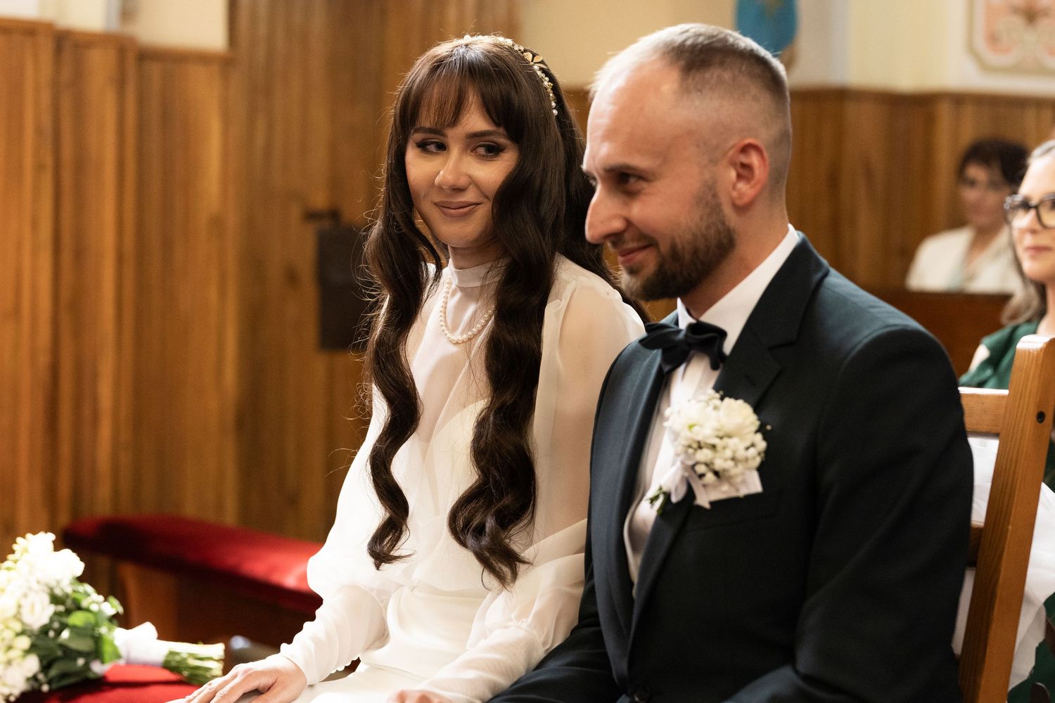 A couple sits together during a wedding ceremony in a wood-paneled church interior, with white floral decorations visible.