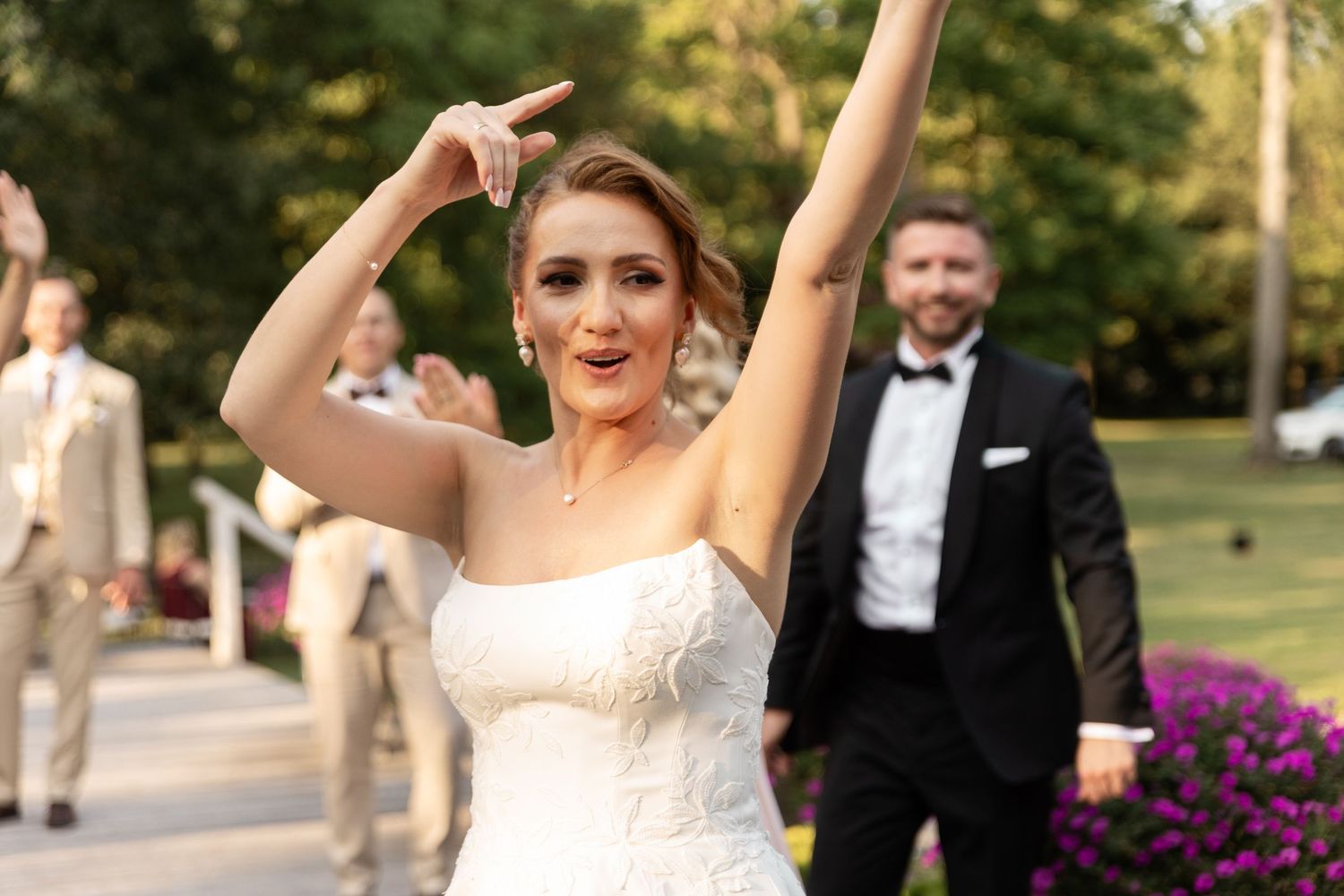 Wedding party members celebrate joyfully outdoors during sunset with raised arms and formal attire.